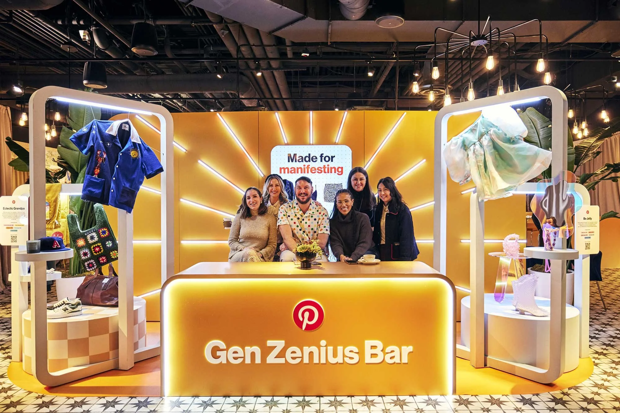 Group of six people sitting and standing behind a yellow Pinterest Gen Zenius Bar booth with clothing and accessories on display, illuminated by warm lighting.