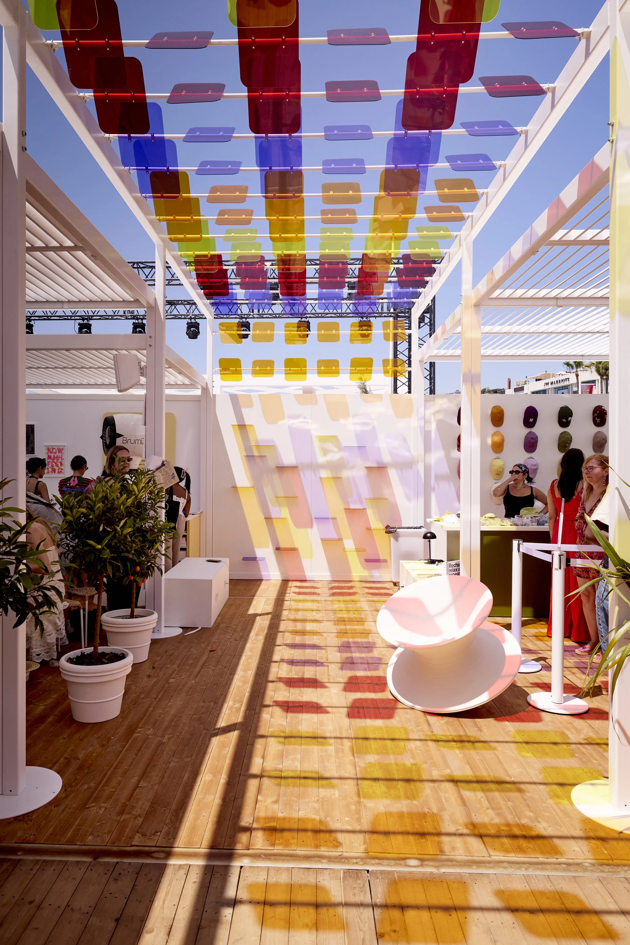 Inside a colorful outdoor art installation with hanging panels casting multicolored shadows on a wooden floor, with people browsing and talking, and a variety of colorful hats displayed on the wall.