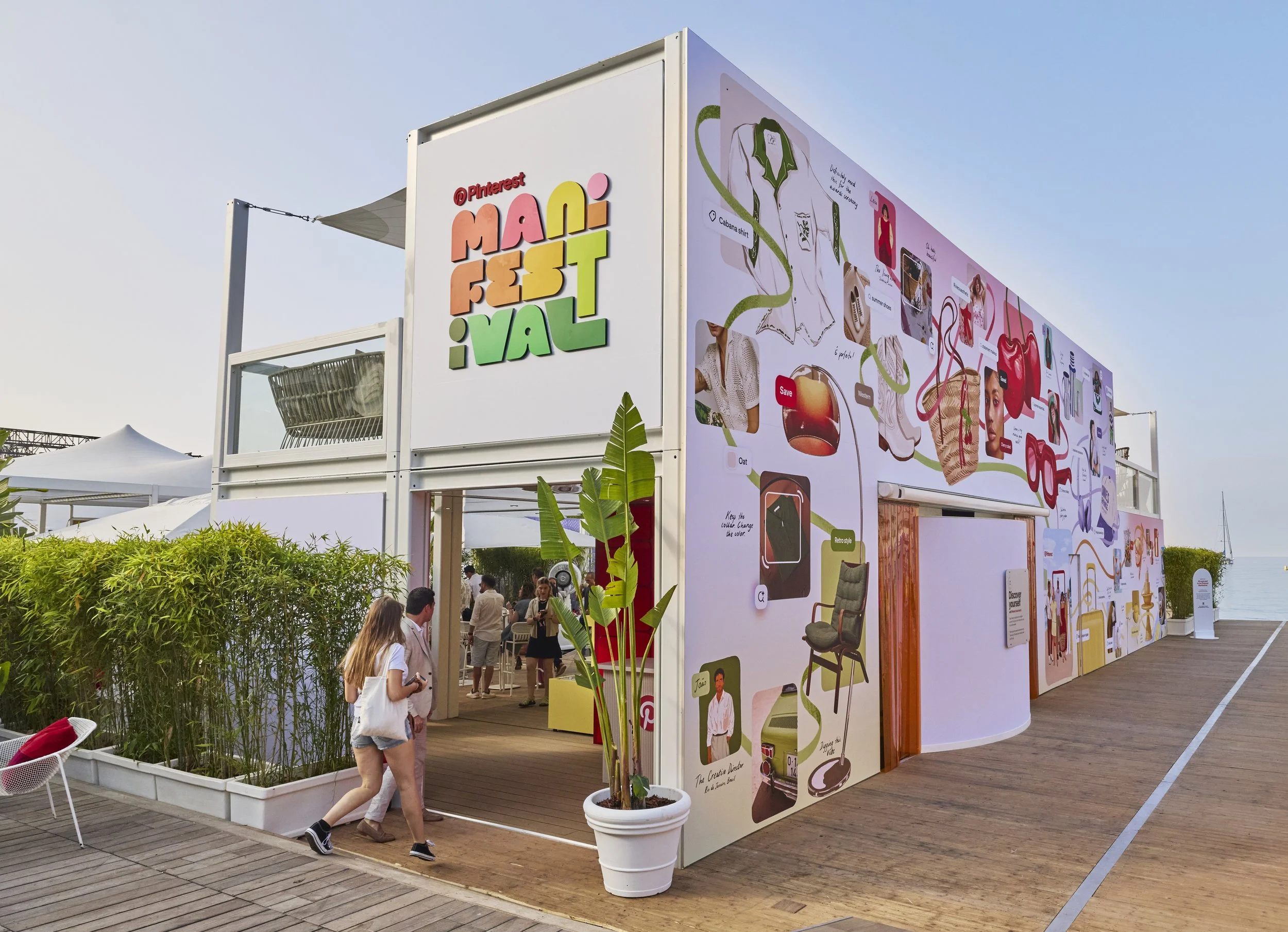 Exterior of a two-story pavilion with a colorful sign reading 'Pinterest Mania Festival' on the front. The building features a vibrant mural with illustrations of various products and objects. Visitors walk on a wooden deck, some entering or exiting 
