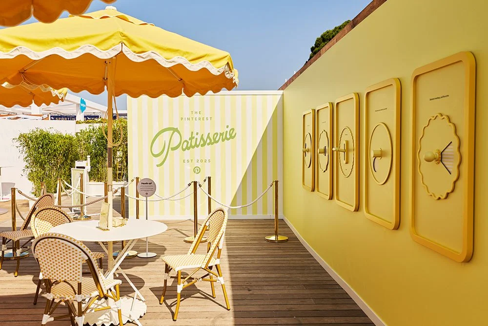 Outdoor patio of a patisserie with yellow decor and striped wall, featuring tables and chairs under yellow umbrellas, with a bright sky overhead.