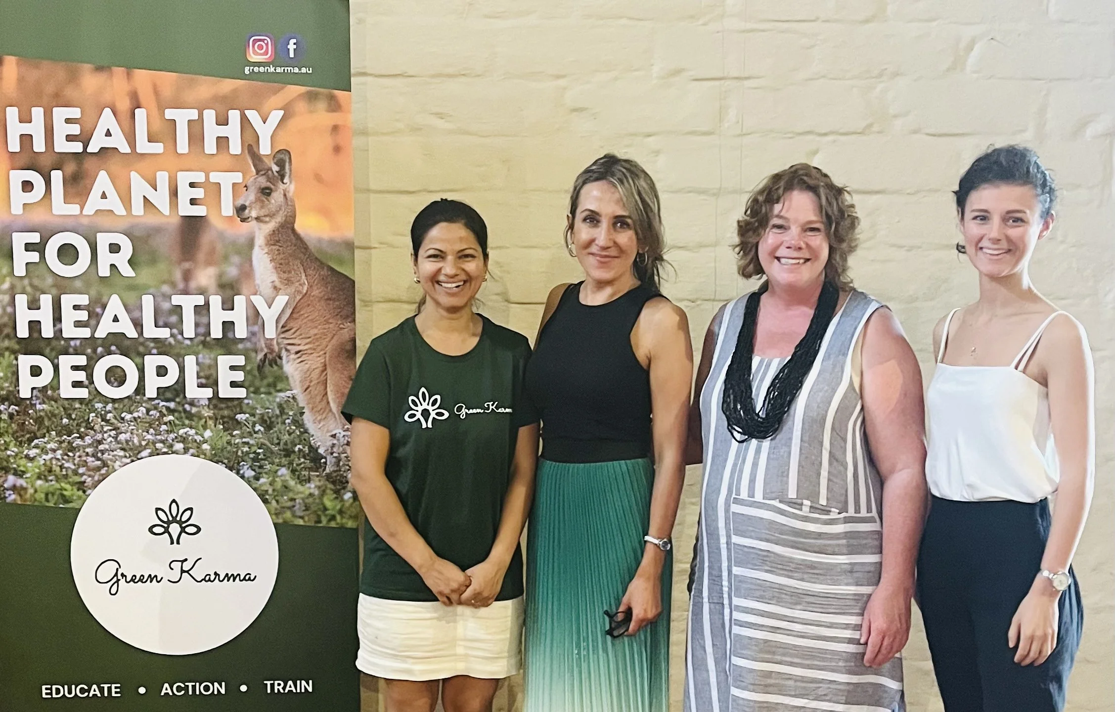 Four women standing next to a Green Karma charity poster that says 'Healthy Planet for Healthy People.' The women are smiling and posing for the photo in front of a beige brick wall.