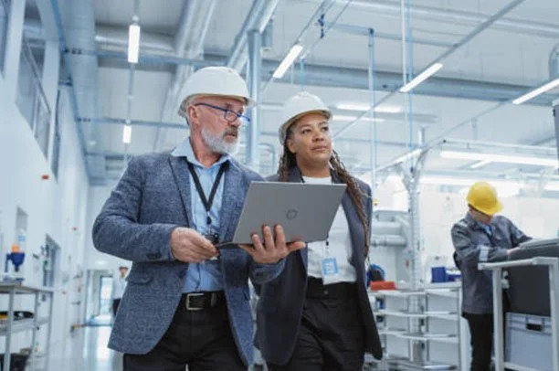 Homens e mulheres usando capacetes de segurança e roupas formais, inspecionando uma fábrica ou planta industrial moderna.