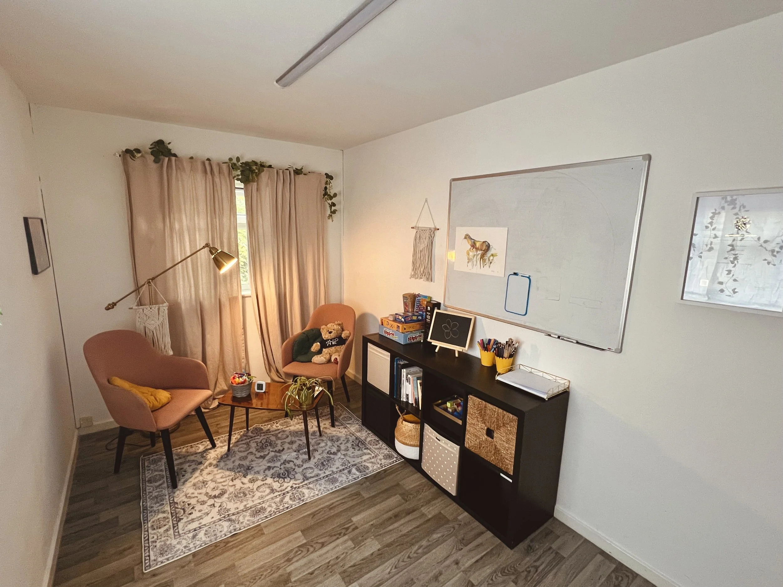 A cozy room with two pink armchairs, one with a yellow blanket, a small wooden coffee table with a plant, a basket, and snacks. Behind the chairs is a window with beige curtains, a gold reading lamp, and a white wall with a framed picture. To the rig