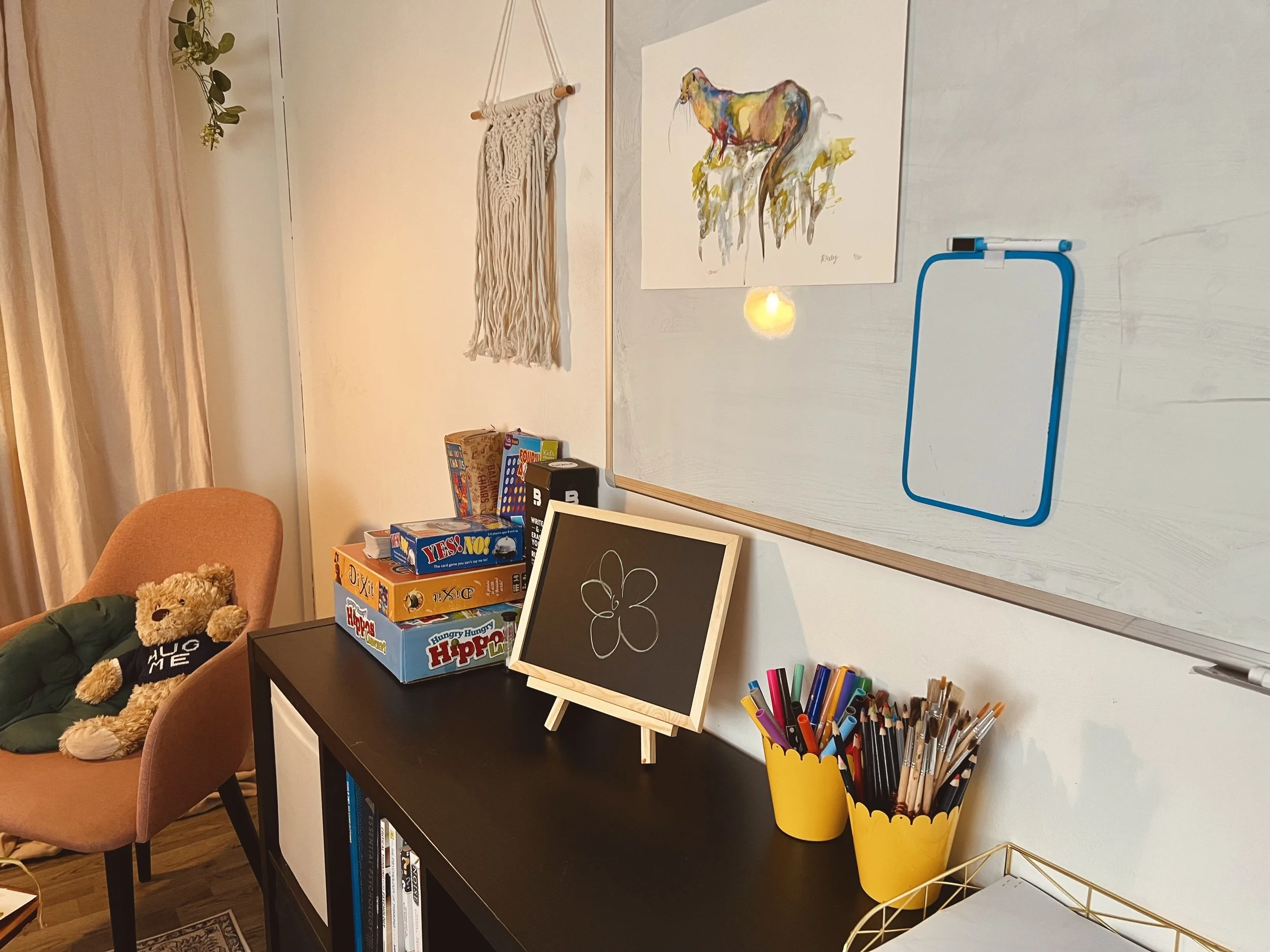 A cozy room corner features a whiteboard, a framed chalkboard drawing of a flower, colorful markers in yellow containers, a stack of board games, a tan armchair with a teddy bear- at Ana Hristov Therapy in Harborne, Birmingham.