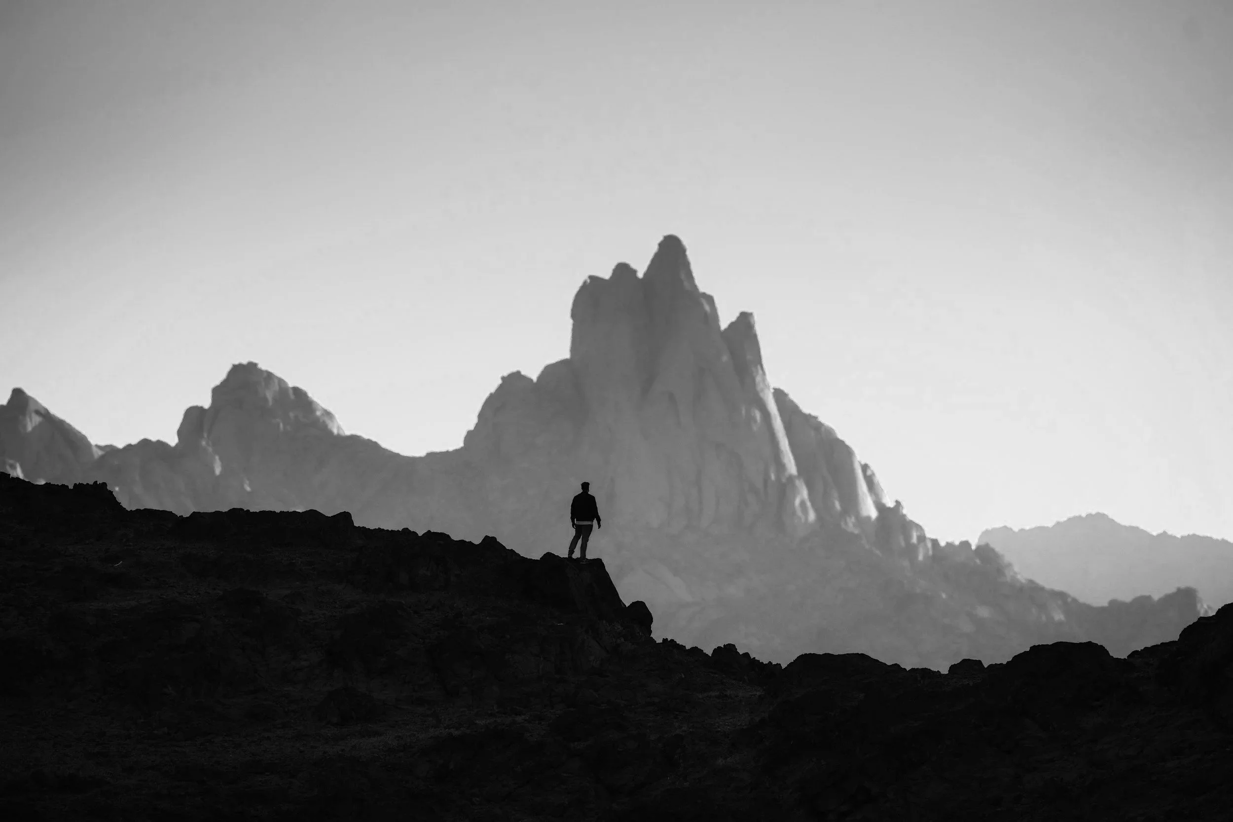 A silhouette of a person standing on a rocky terrain with large mountain peaks in the background, in a black and white landscape photograph.