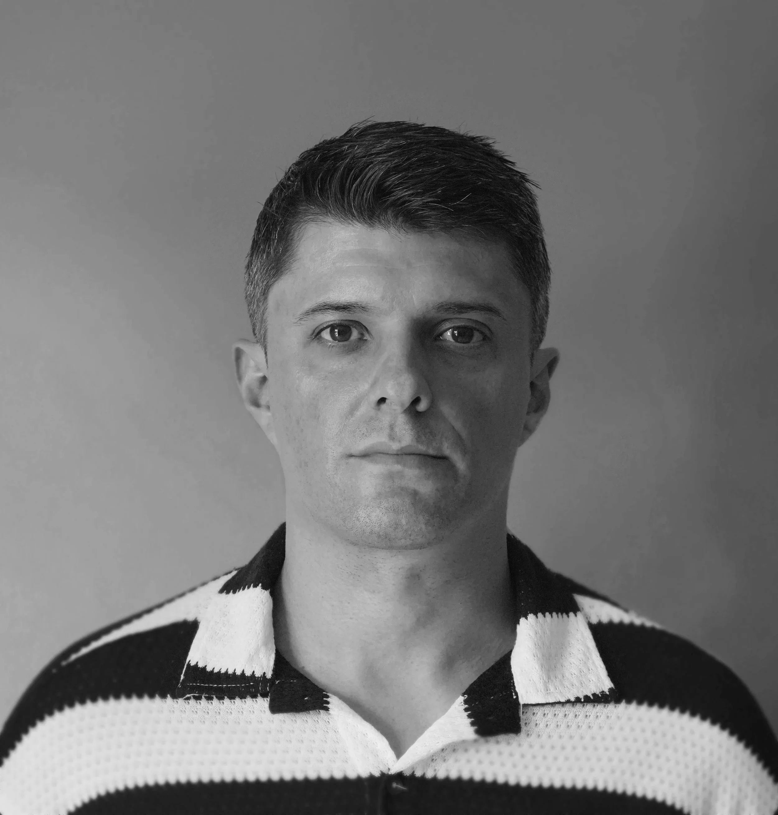 A black-and-white portrait of a man with short, dark hair, wearing a striped polo shirt, looking directly at the camera against a plain background.