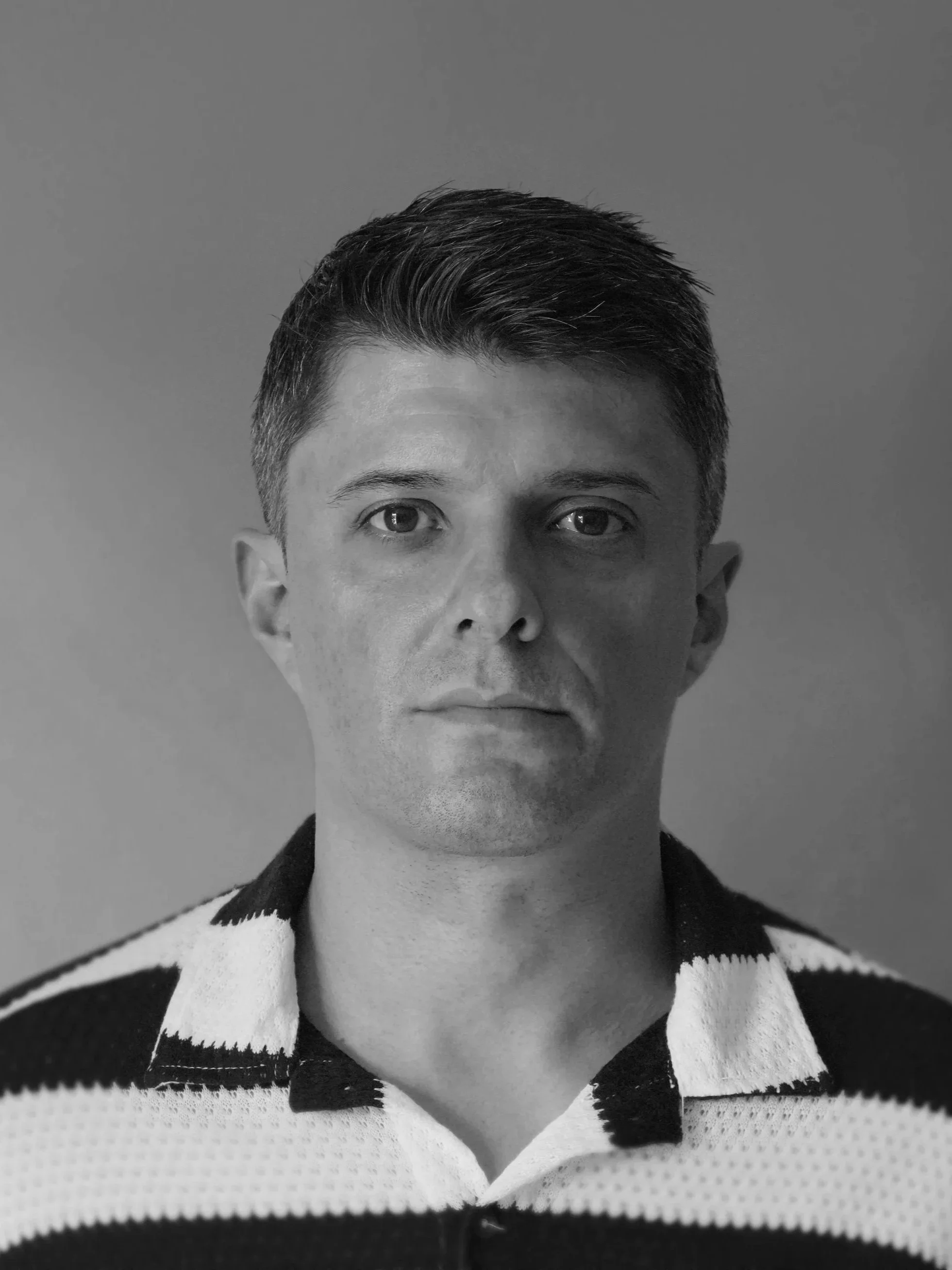 A black and white portrait of a man with short, dark hair, wearing a collared, horizontally striped shirt.