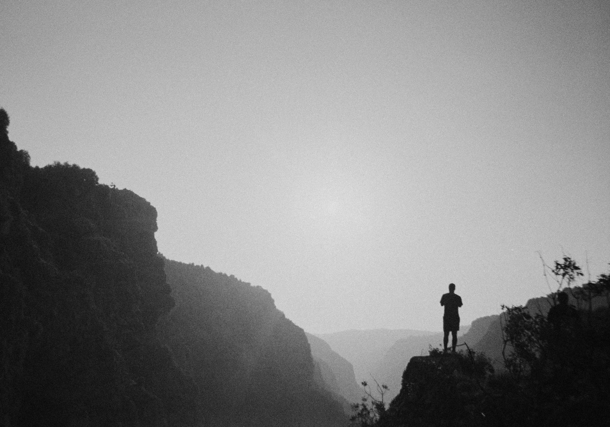 Silhouette of a person standing on a rocky ledge overlooking a valley with cliffs, in black and white.