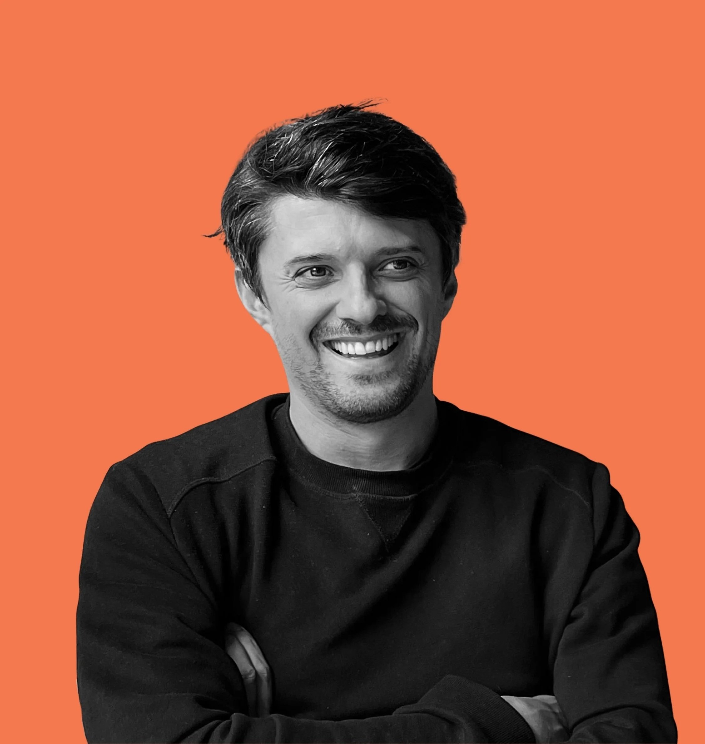 A smiling man with dark hair and a beard, wearing a black sweatshirt, sitting with his arms crossed against an orange background.