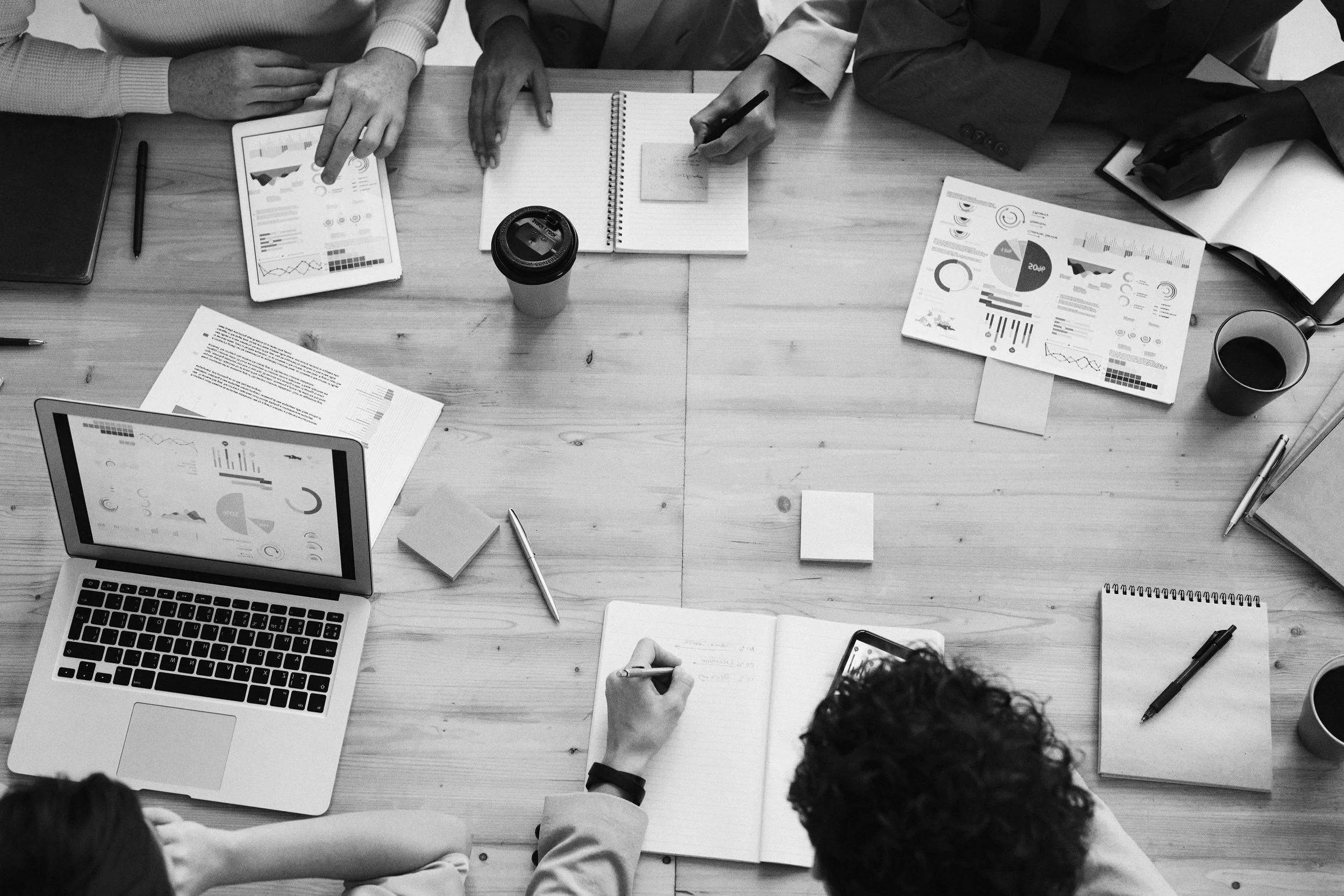 People sitting around a wooden table working on laptops, notebooks, and printed charts, with coffee cups and stationery.
