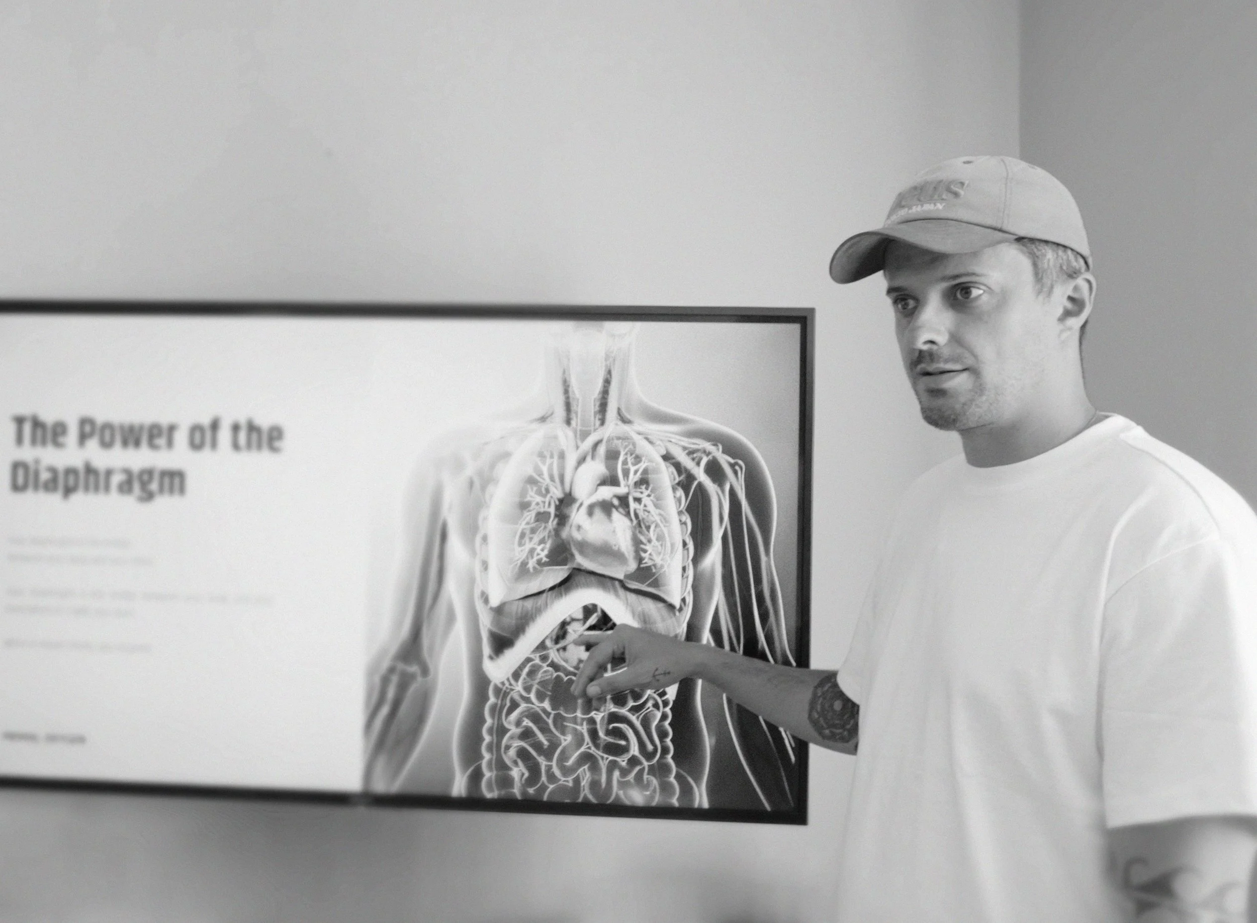 A man in a baseball cap and white t-shirt pointing at an anatomical diagram of the human body on a screen, titled 'The Power of the Diaphragm', showing internal organs and nerves.