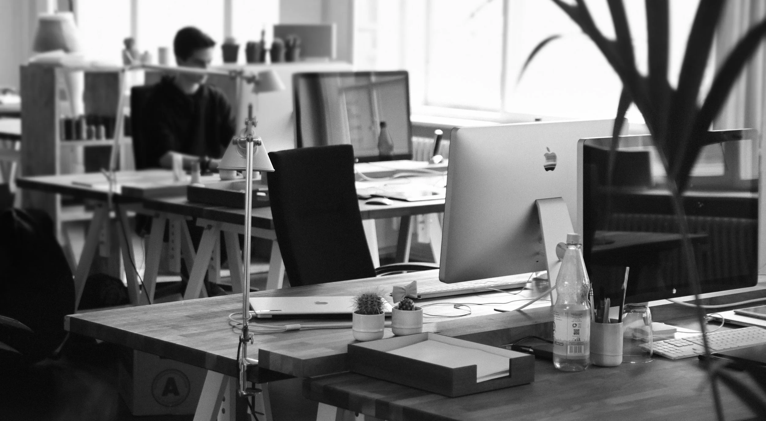 Office workspace with desks, computers, and office supplies, and a person working in the background.