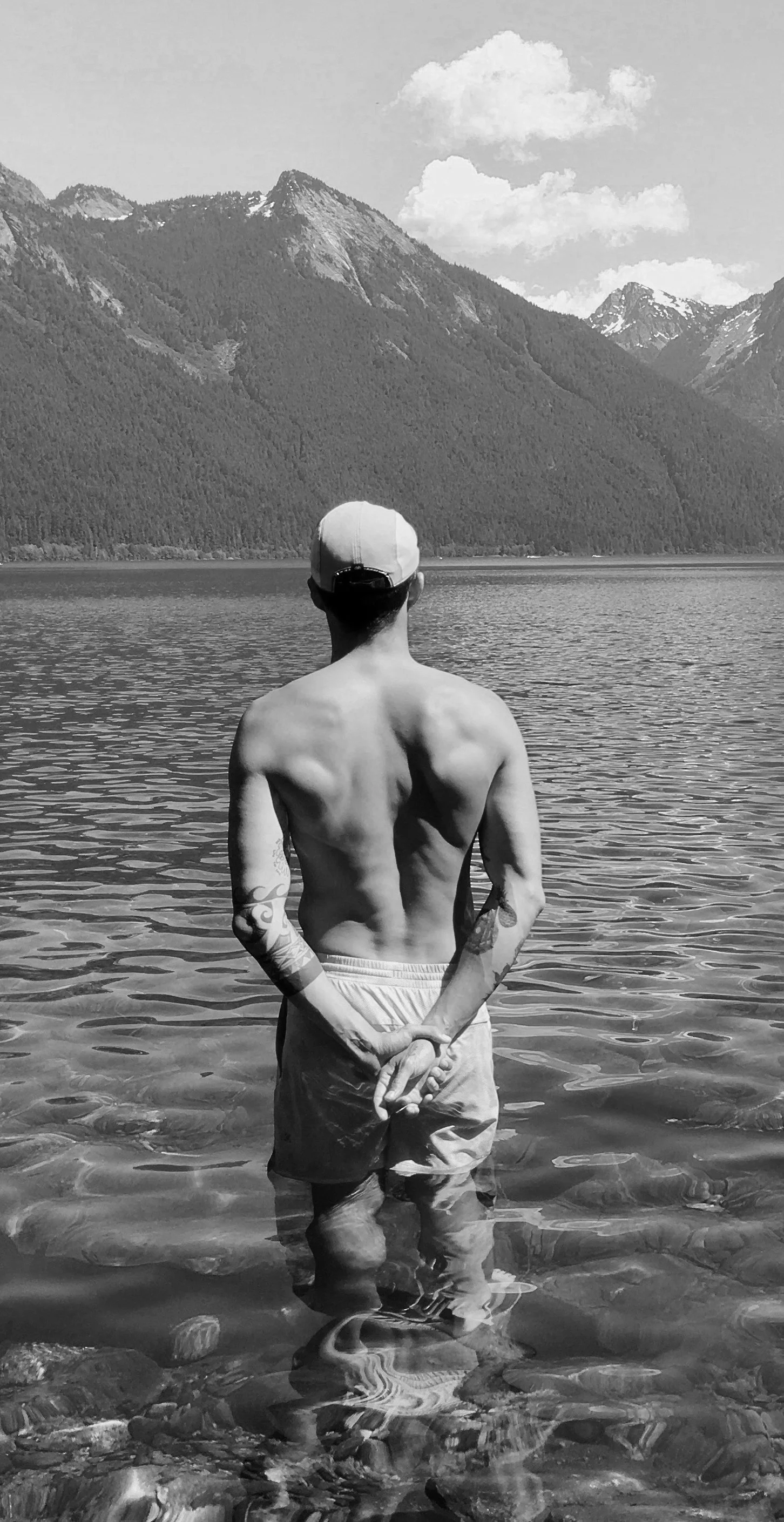 A shirtless man with tattoos on his arms stands in a lake, facing away from the camera towards mountains and clouds in the distance, wearing shorts and a baseball cap.