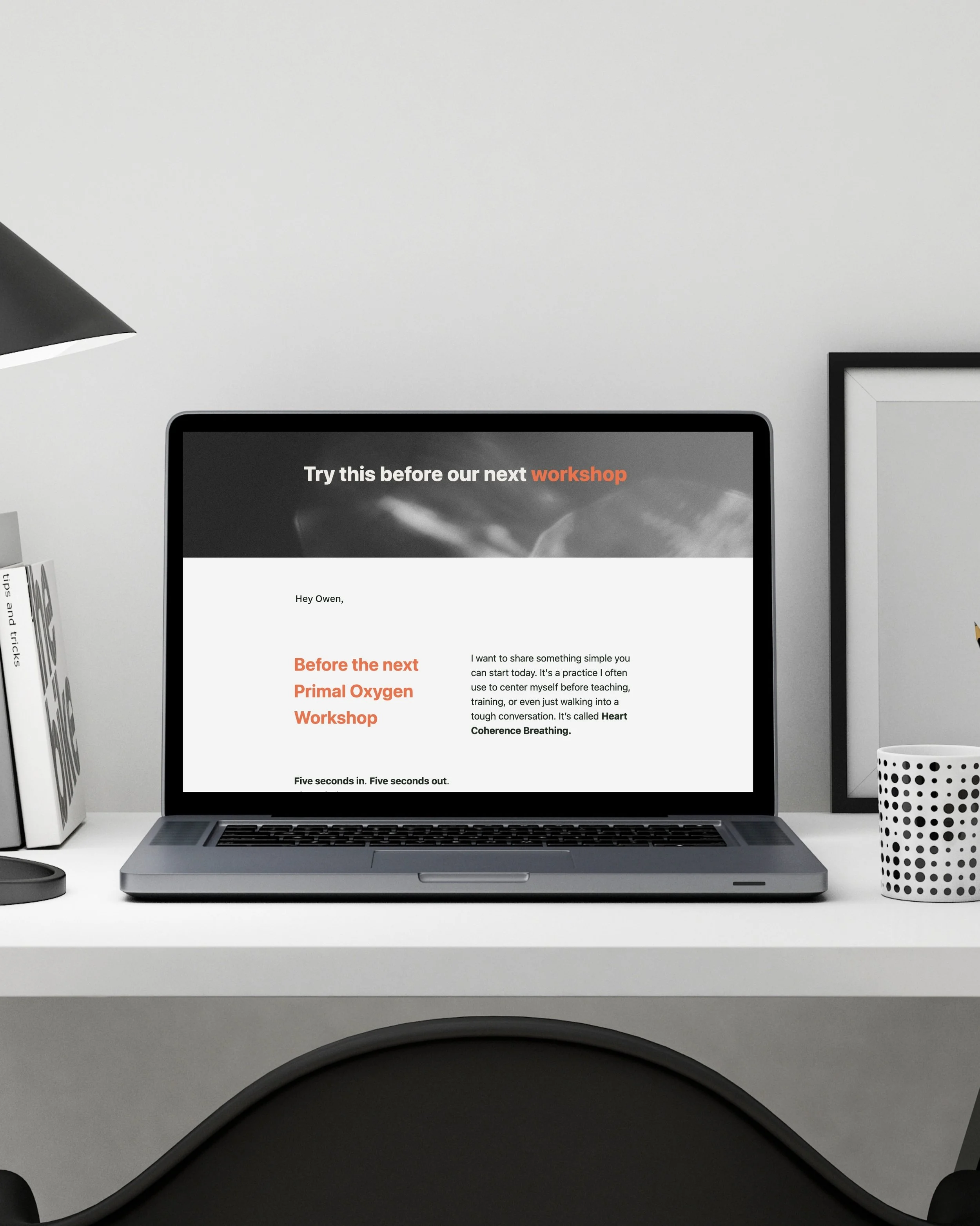 Laptop on white desk displaying a digital page about a Primal Oxygen Workshop, with a black desk lamp, a polka-dotted cup, and a framed picture in the background.