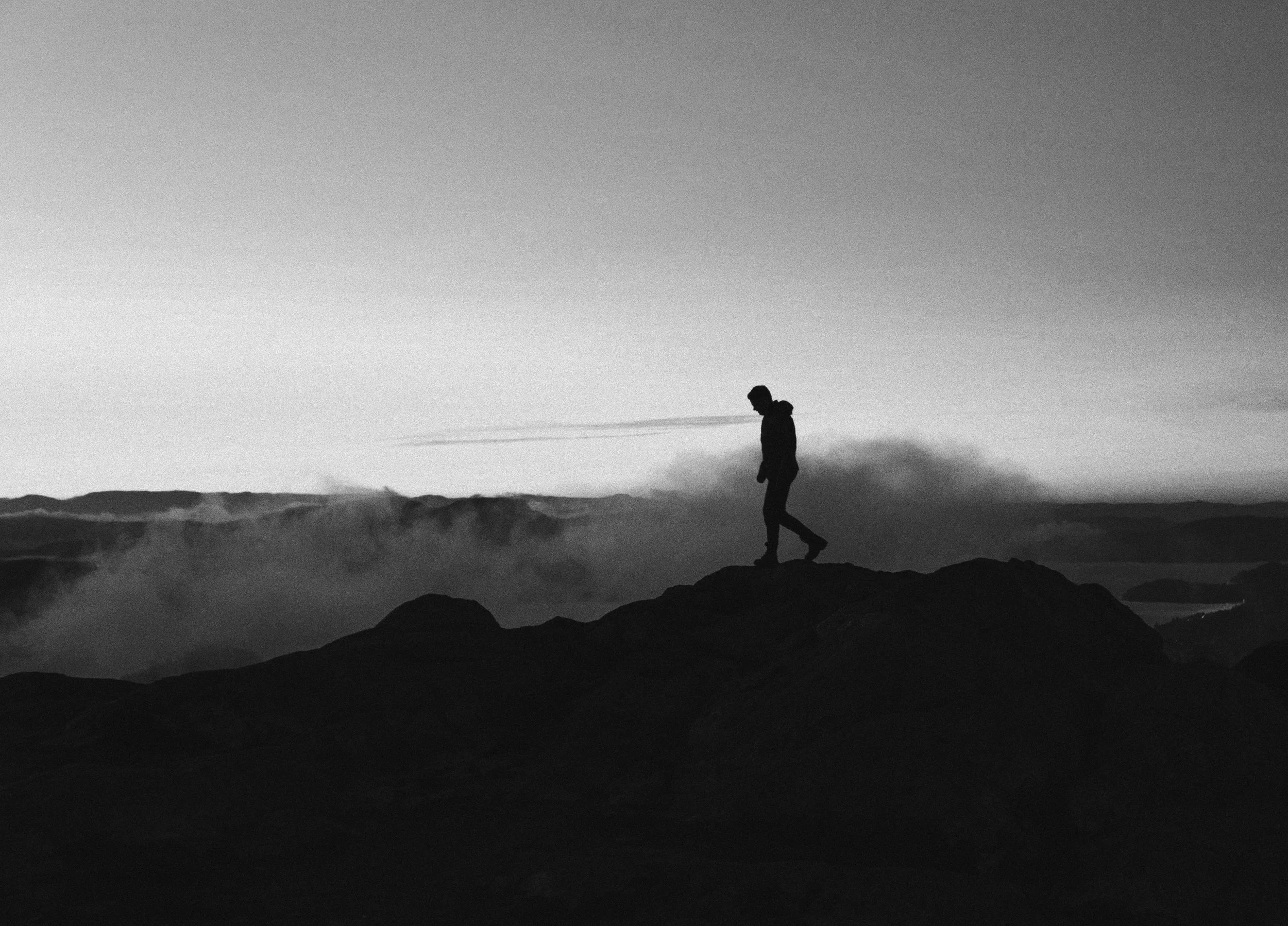 A black and white silhouette of a person standing on top of a mountain, overlooking a cloudy landscape and distant mountains during sunset or sunrise.