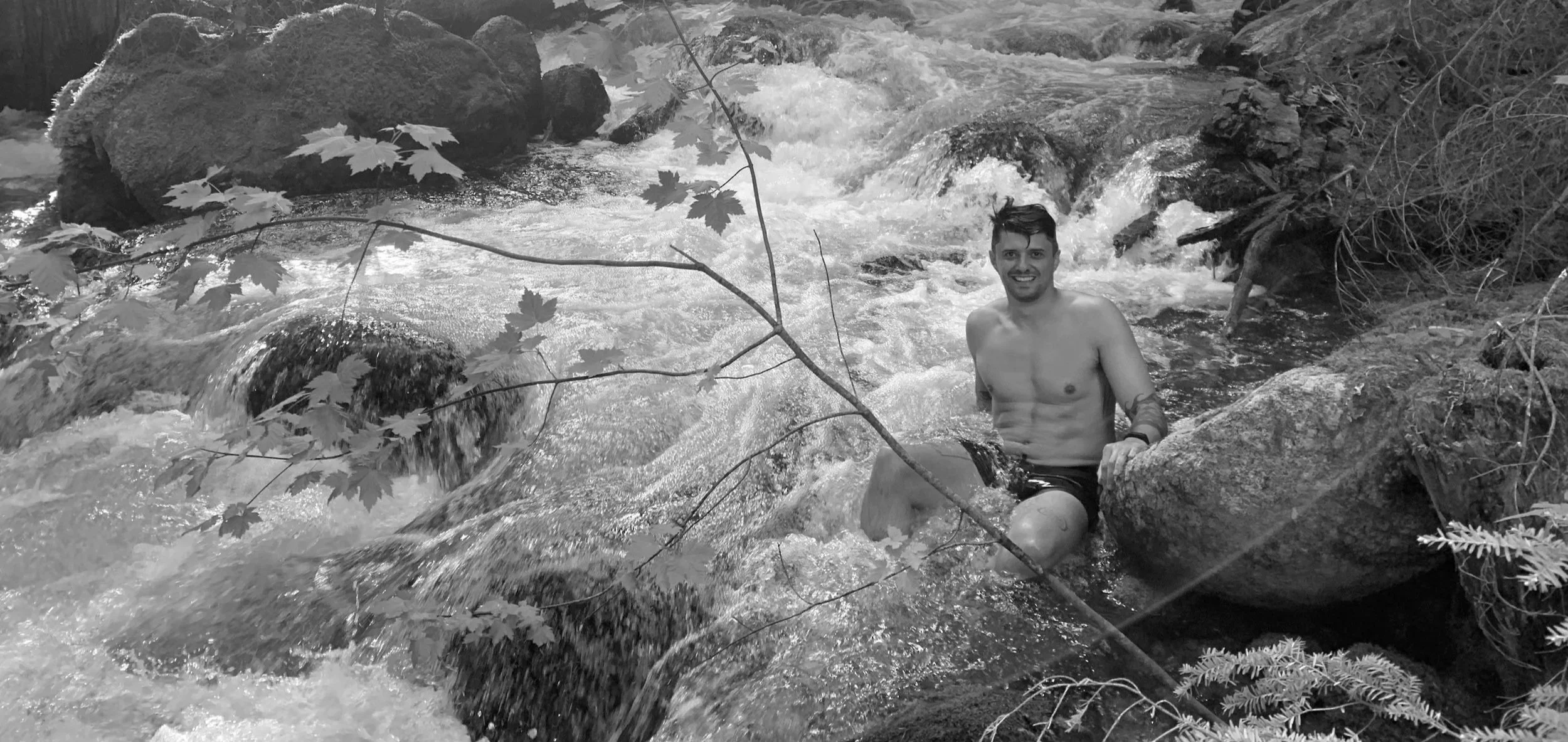 A smiling shirtless man sitting on a large rock in a rushing river surrounded by rocks and trees.