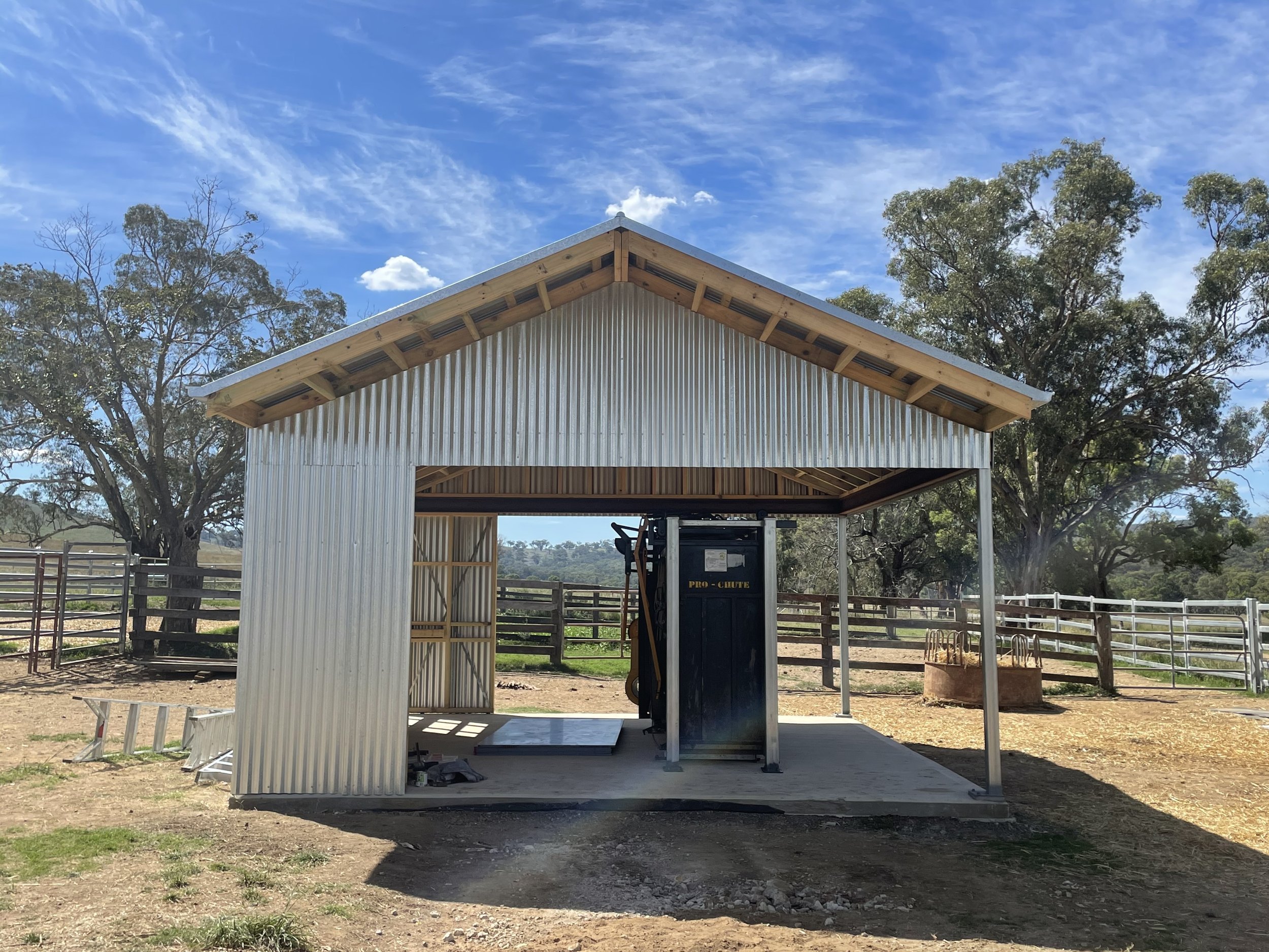 A partially constructed metal shed in a rural area with trees and fences in the background under a partly cloudy sky. Farm shed in Mudgee