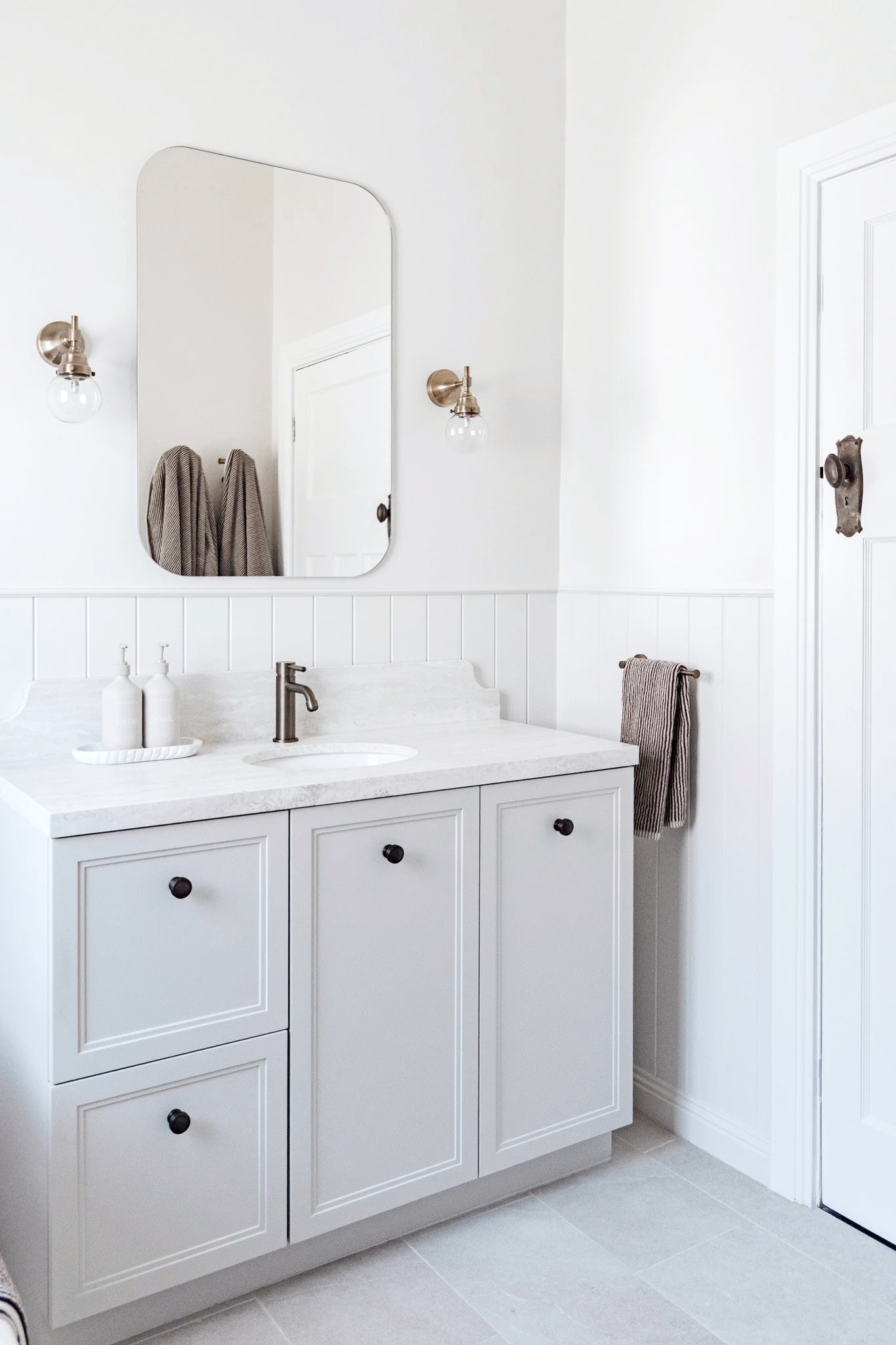 Bright bathroom vanity with white cabinet, marble countertop, and oval mirror. Wall-mounted light fixtures with exposed bulbs, grey towels hanging, and minimal decor. Bathroom Renovations Mudgee