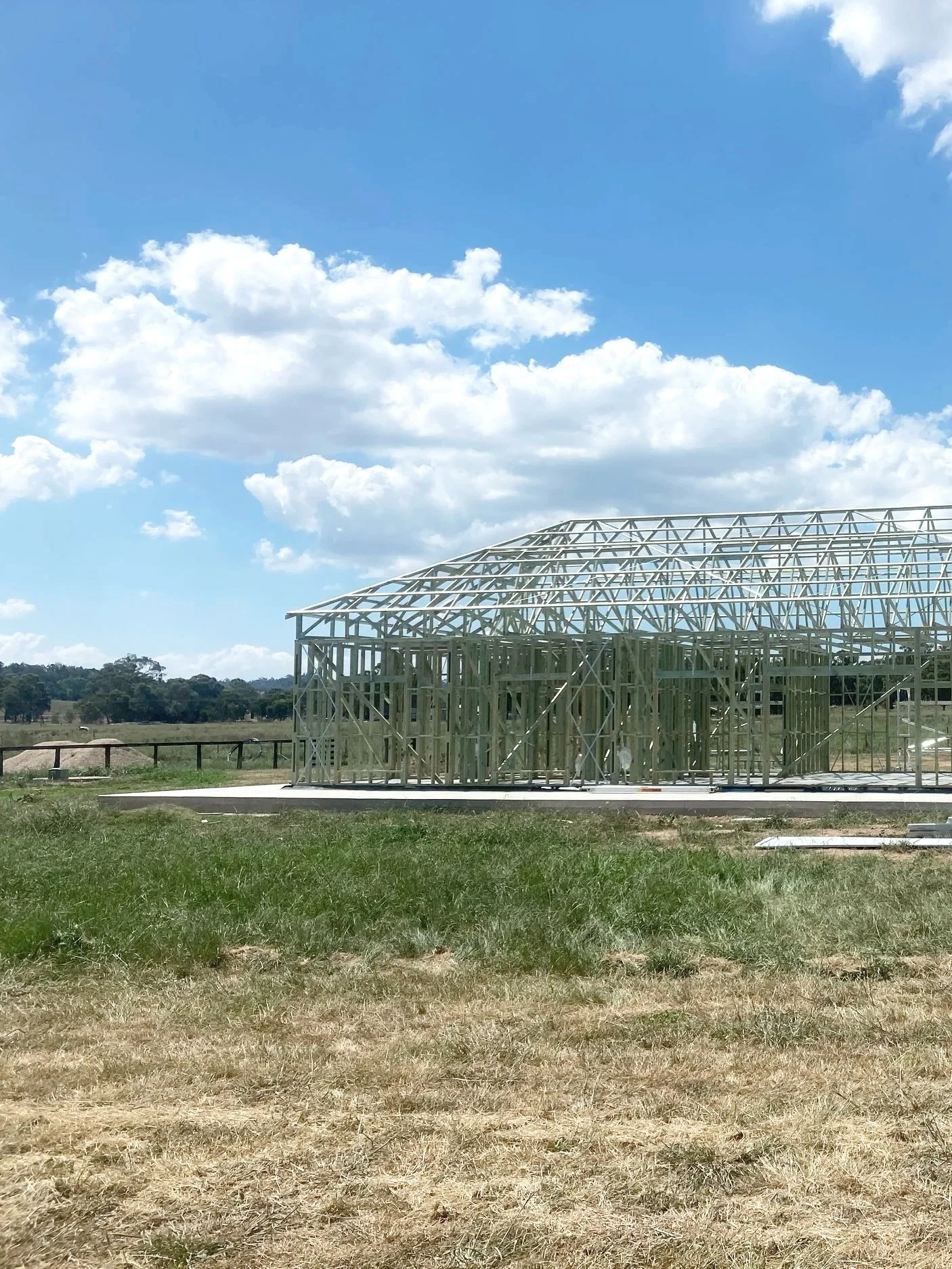 A wooden house frame under construction outdoors with a grassy field, a fence, trees in the background, and a partly cloudy blue sky. Located in Mudgee NSW