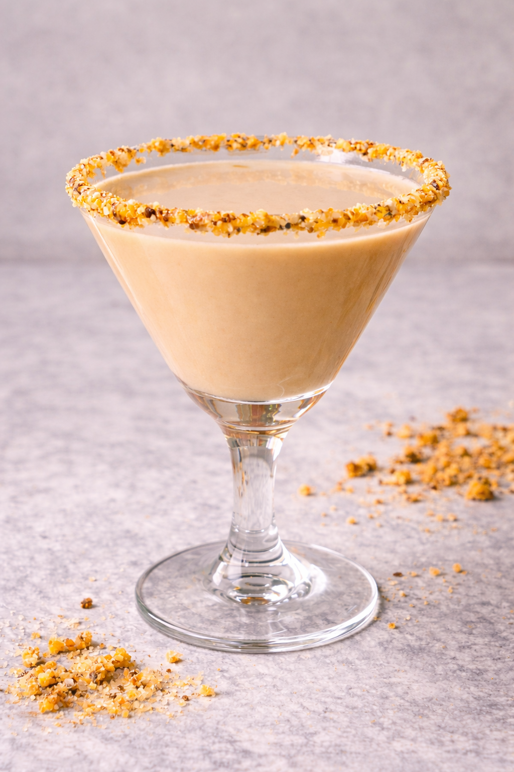 A creamy, light brown cocktail in a martini glass with a crushed nut rim, placed on a gray surface with some crushed nuts scattered around.