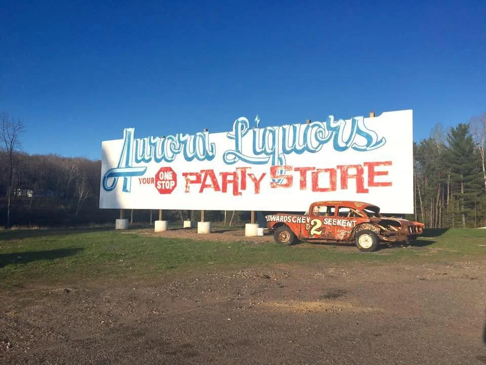 Large billboard for Aurora Liquors, offering one-stop party store, with a vintage race car parked in front.