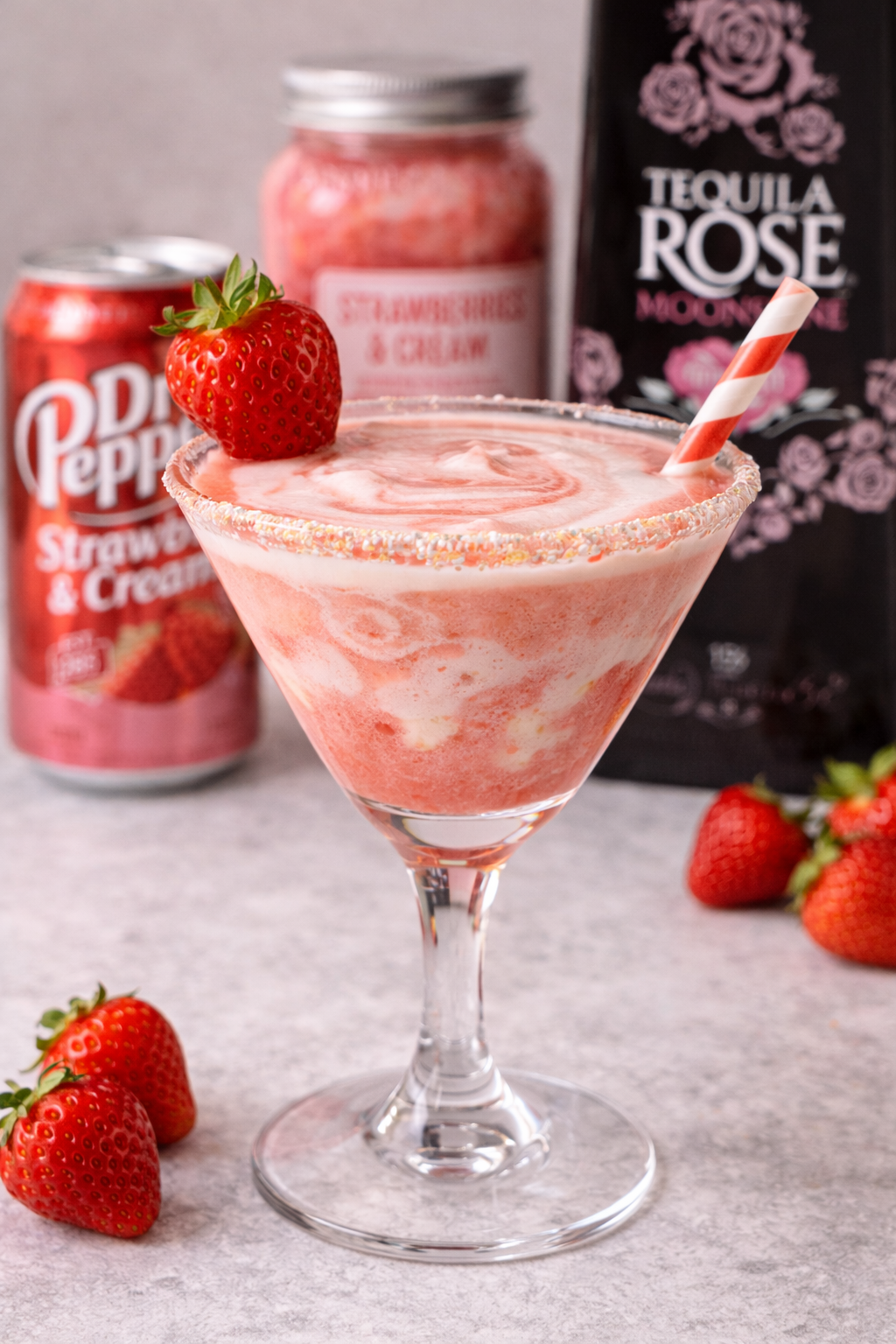 A strawberry frozen drink in a cocktail glass with a strawberry on the rim and a striped paper straw. Background includes a can of strawberry & cream drink, a jar of strawberries & cream, and a black bag of Tequila Rose.
