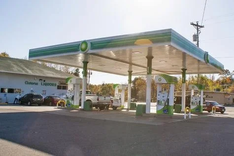 A gas station with green and white branding, fuel pumps, and several parked cars, located in a sunny outdoor area.
