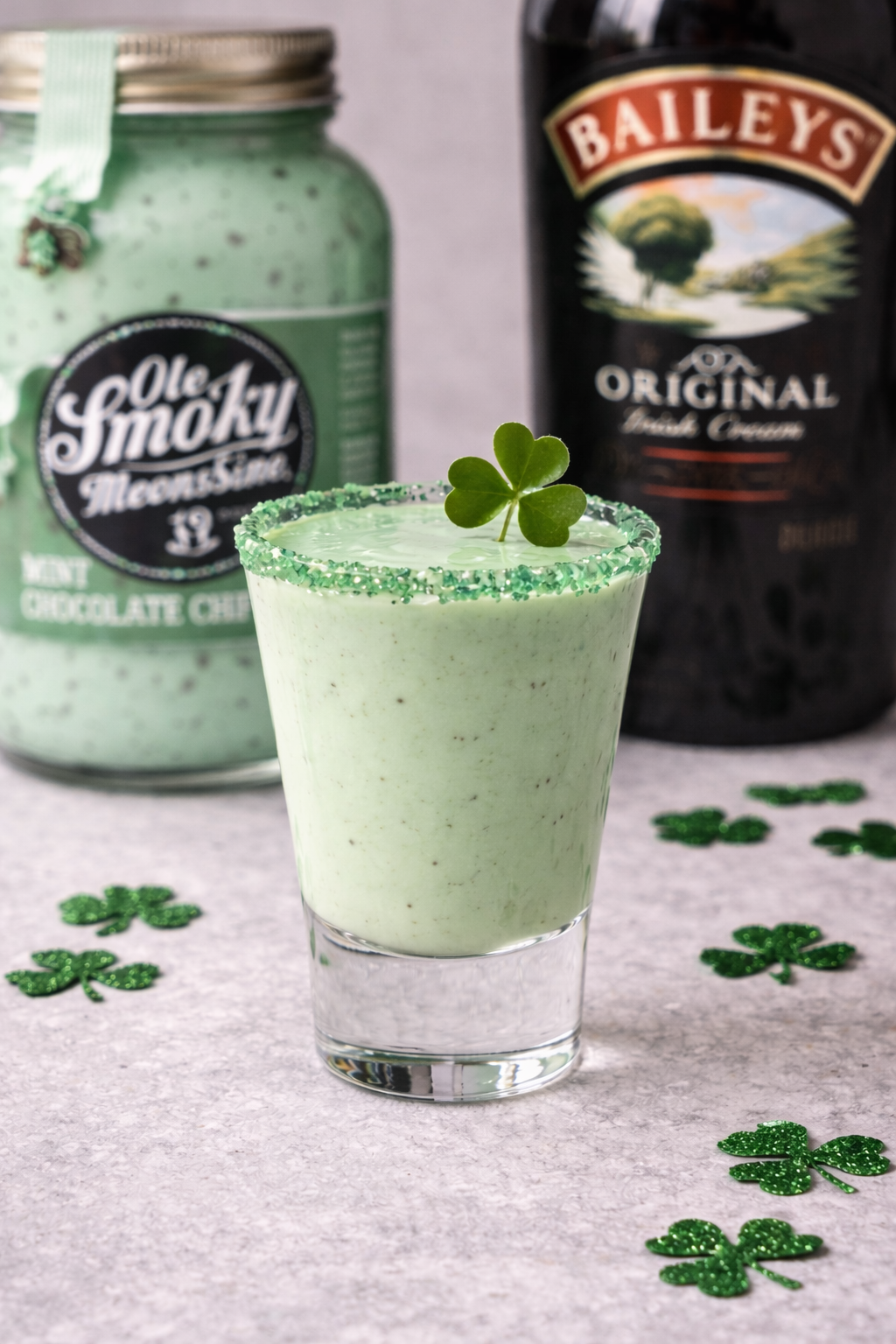 A green Shamrock-themed milkshake garnished with a shamrock leaf and green sprinkles, with containers of Bailey's Irish Cream and chocolate chip ice cream in the background, surrounded by glittery shamrock decorations.