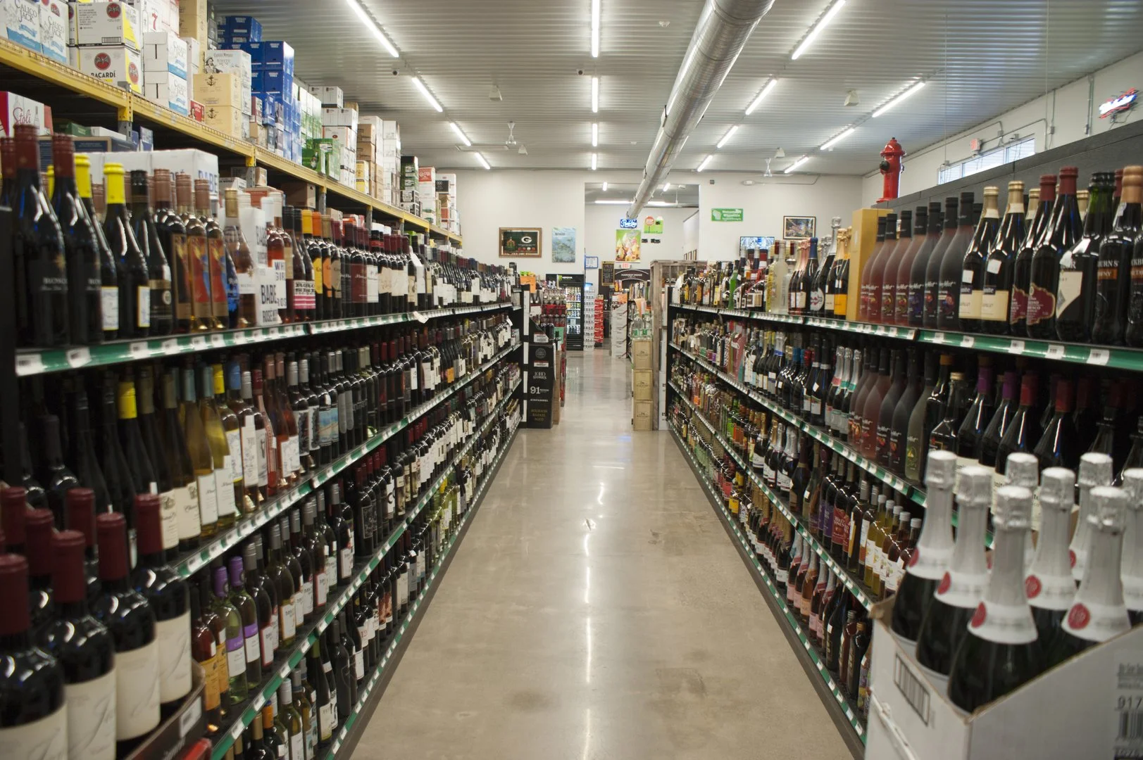 Supermarket aisle with shelves stocked with wine bottles, beer bottles, and other alcoholic beverages, with bright overhead lighting and a wide walkway.
