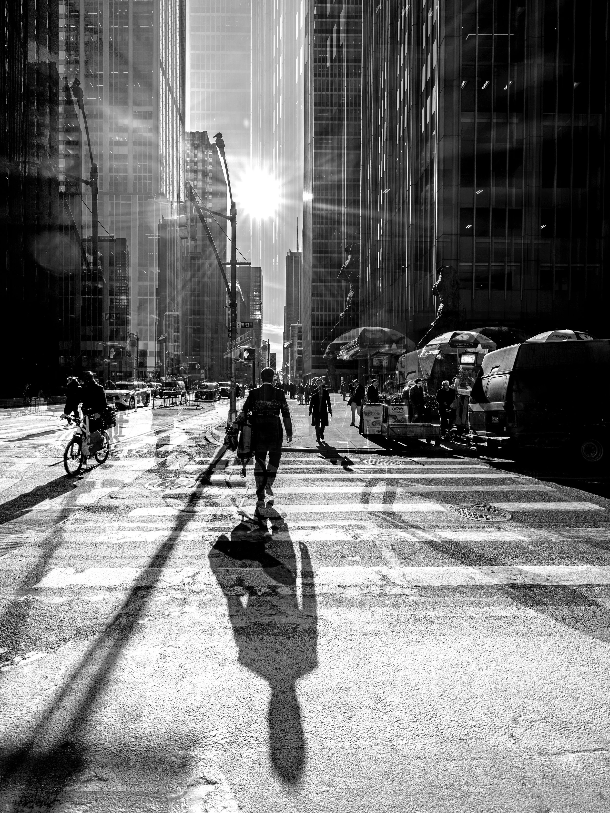Black and white photo of a city street with pedestrians, cyclists, and tall buildings. The sun is shining brightly, casting long shadows.
