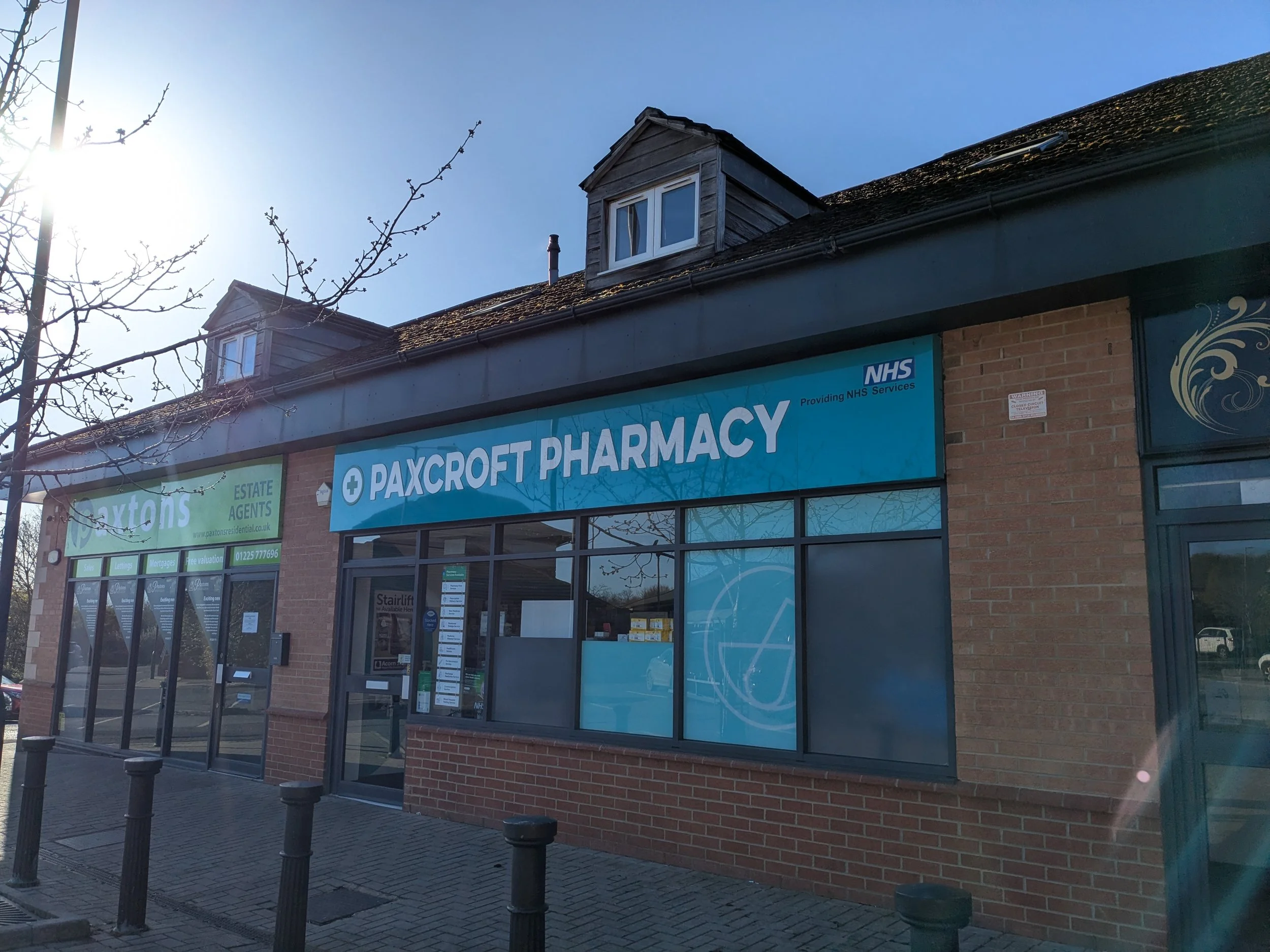 Exterior of Paxcroft Pharmacy with blue signage, large windows, brick walls, and a small parking area with bollards in front, during daytime.