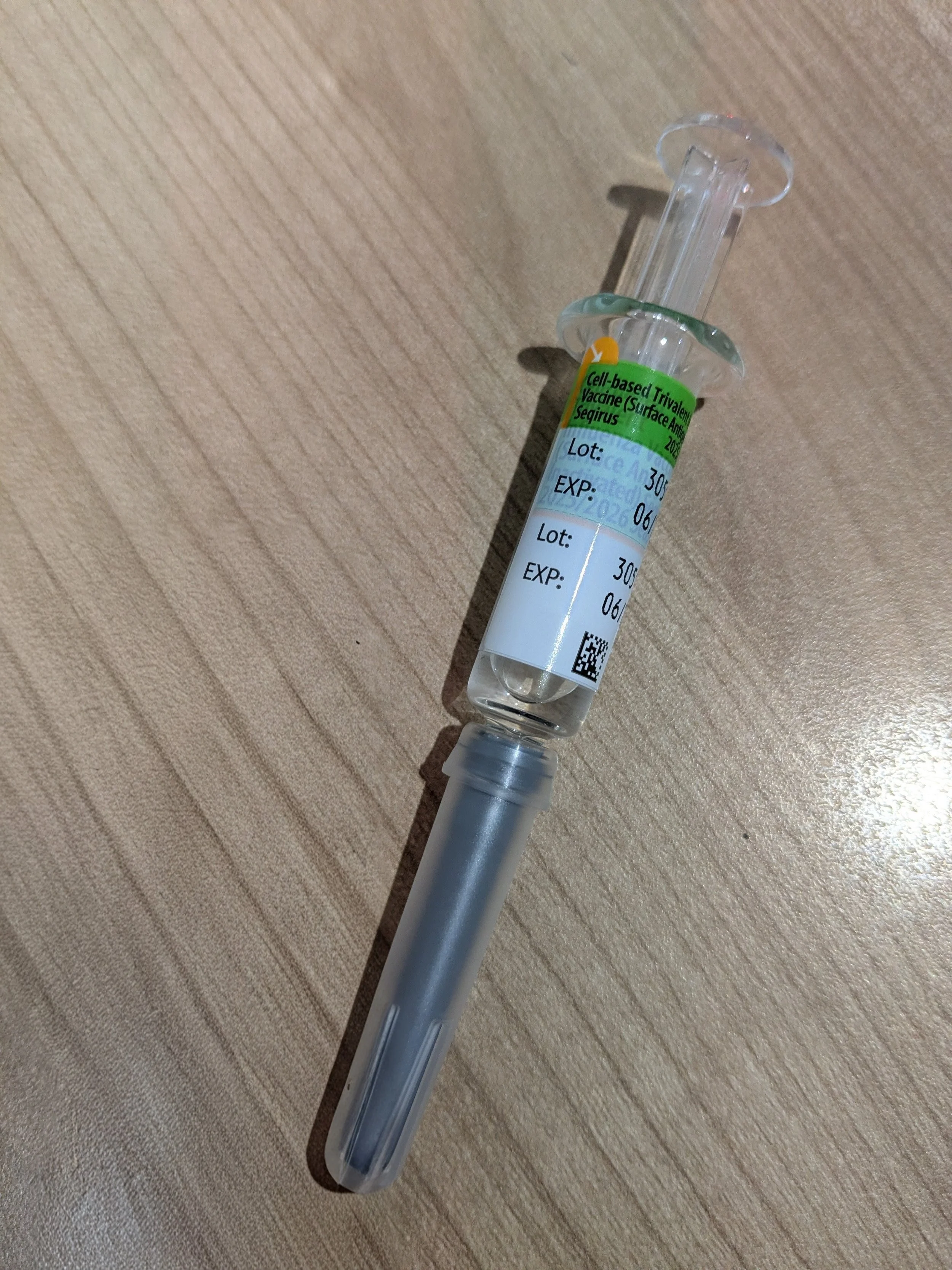A small glass vial with a syringe attached, resting on a wooden surface. The vial has a label indicating it contains a cell-based vaccine.
