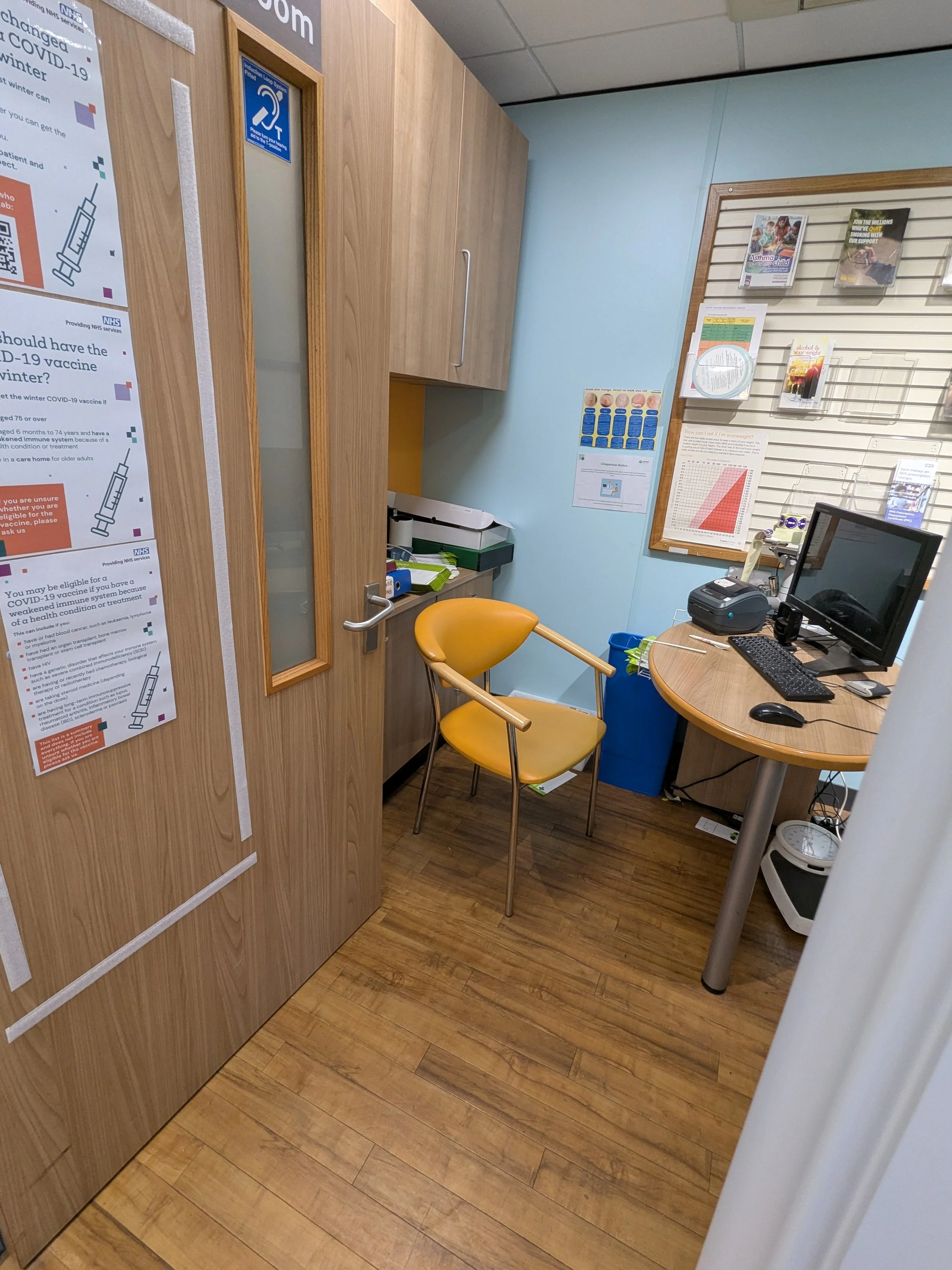 A small medical consultation room with a wooden floor, light blue walls, and wooden furniture. There is a yellow chair with metal arms and a small round wooden desk with a computer, keyboard, mouse, and printer. A large wooden cabinet and a corkboard with magazines and posters are on the wall. A door with frosted glass and posters about COVID-19 vaccination is visible.