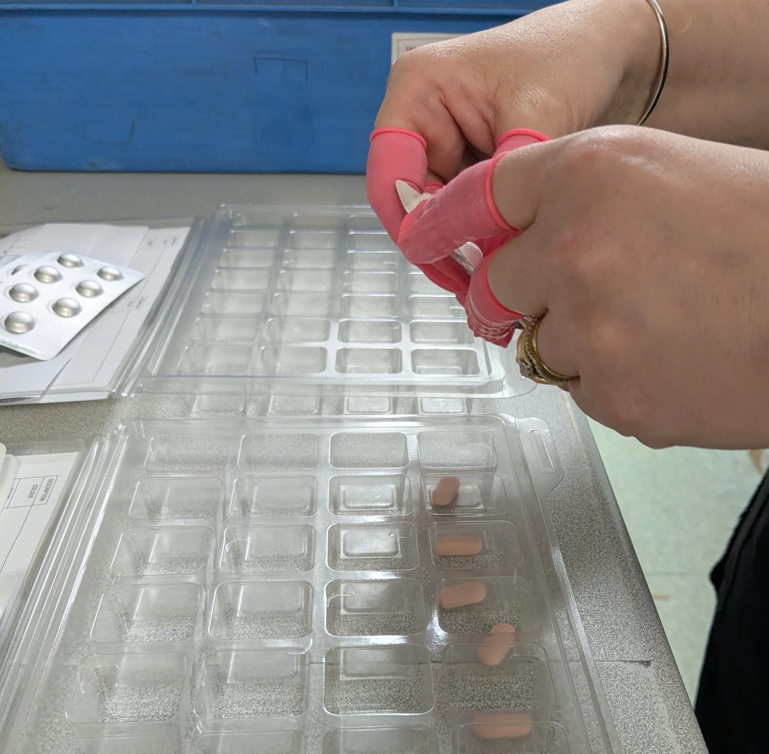 A person wearing pink gloves handling blister packs with pills, preparing to place pills into a plastic container with empty compartments.