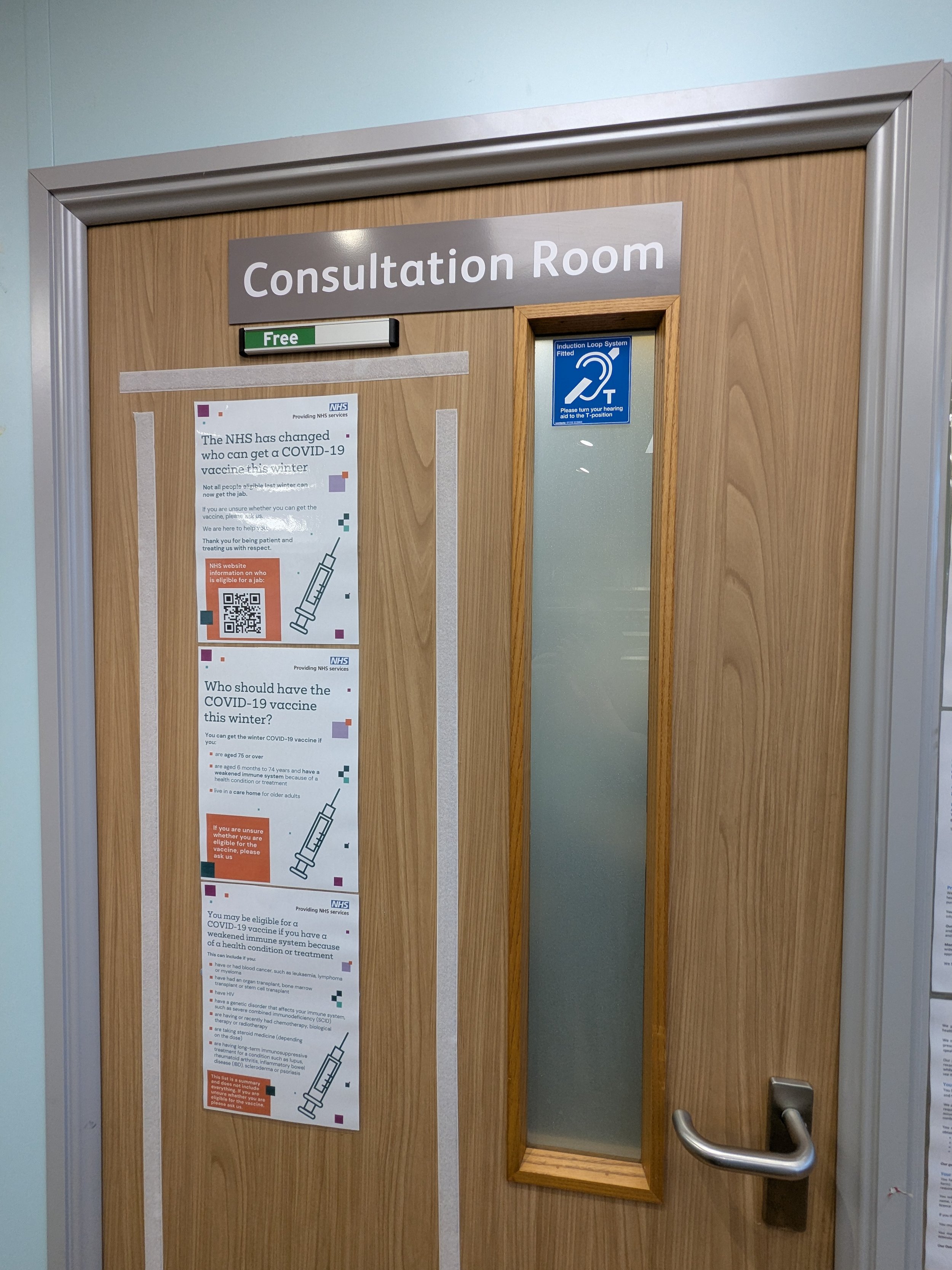 Wooden door with a sign that reads 'Consultation Room' and posters about COVID-19 vaccines posted on it.