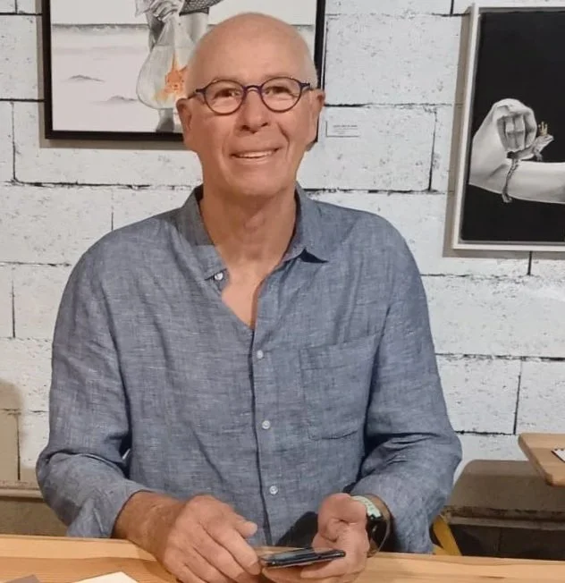 Un homme âgé en chemise bleue, portant des lunettes, souriant, assis à une table dans un espace décoré avec des œuvres d'art sur un mur en bois blanc.