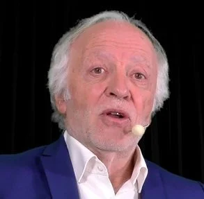 Homme âgé parlant lors d'une conférence, portant un costume bleu et une chemise blanche, avec un micro attaché à l'oreille.
