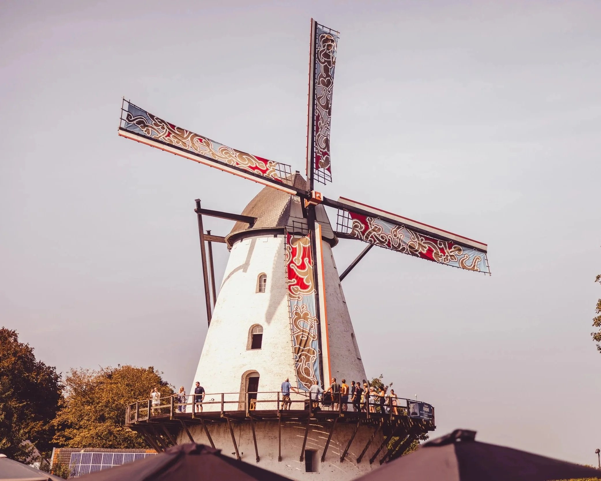 A traditional windmill with blades decorated with colorful patterns, standing against a cloudy sky, with visitors walking around the platform at its base.