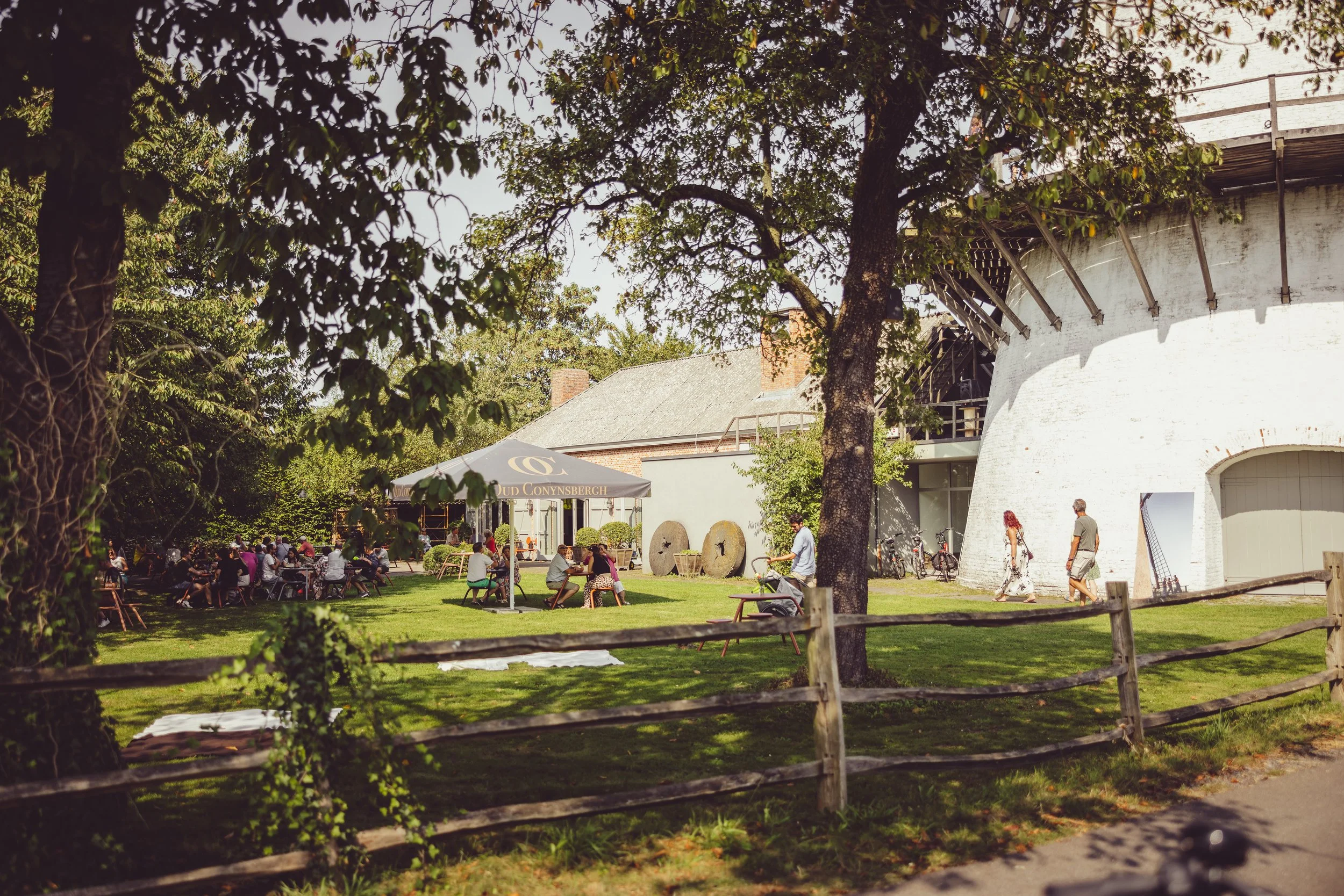 People gathered outdoors at a social event on a sunny day, sitting at tables under umbrellas, with trees and a white round building in the background.