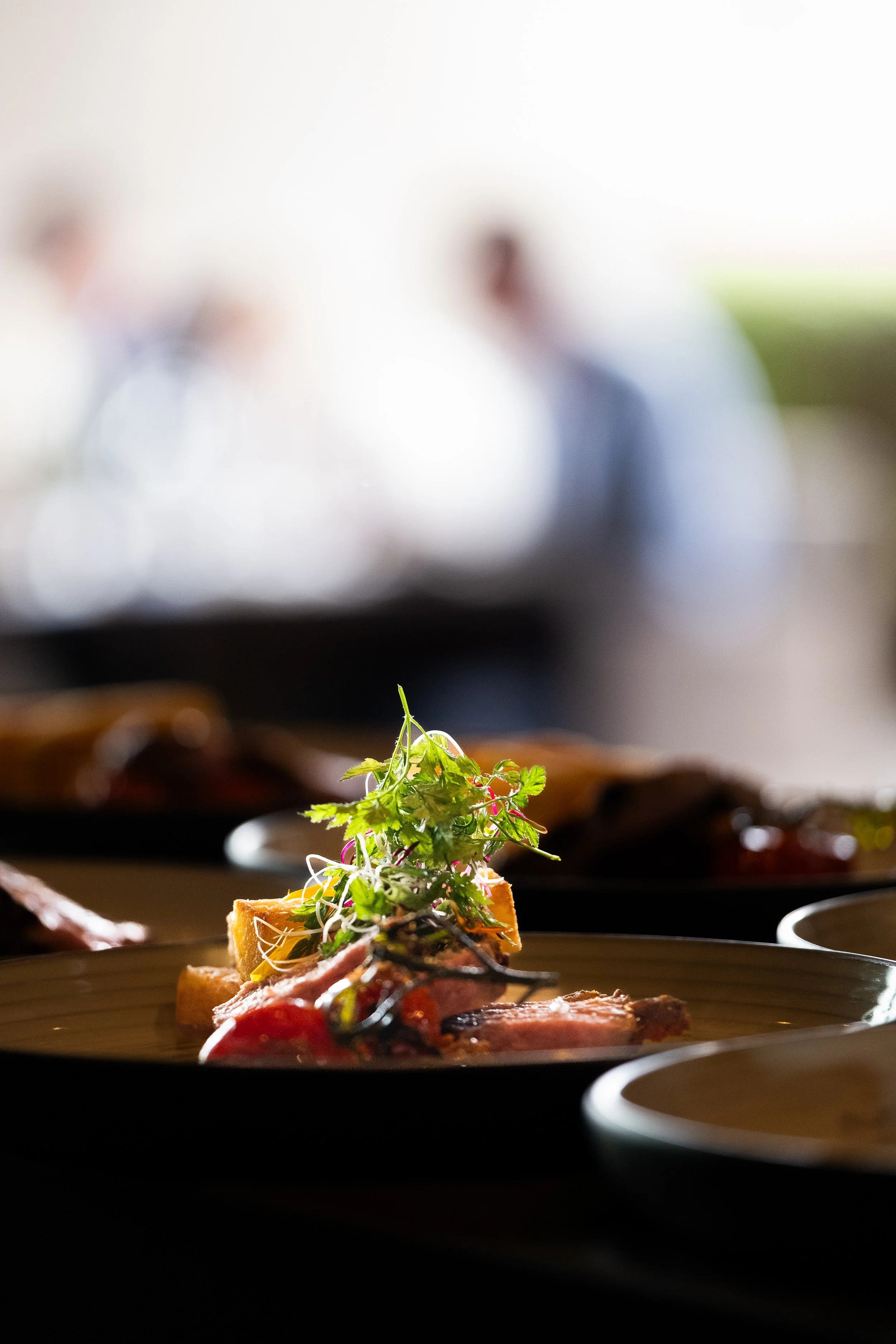 Close-up of a plated gourmet dish with microgreens on top, with a blurred background of two people in a bright restaurant or dining area.