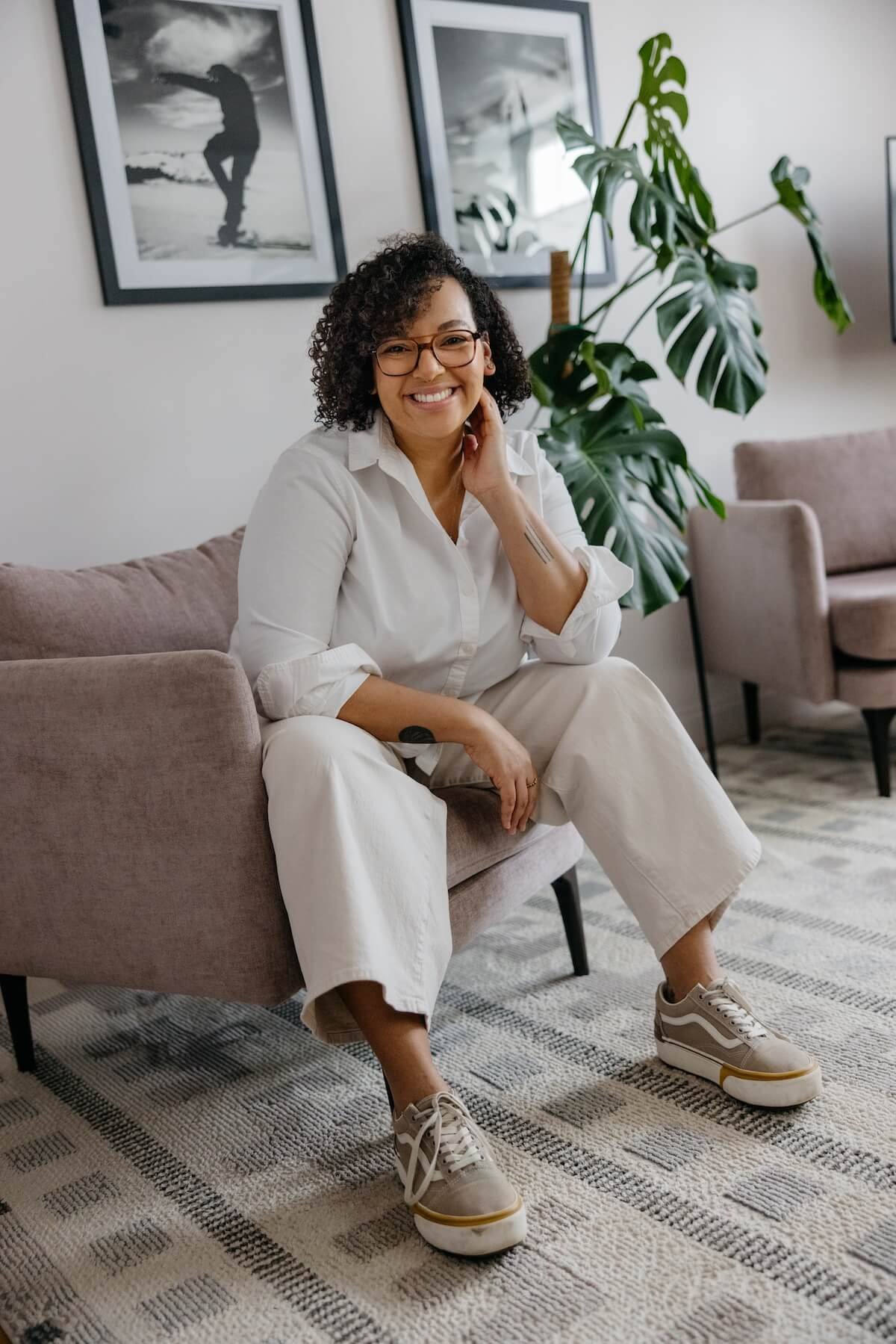 Brittney Nichols, a smiling woman wearing glasses, a white shirt, and beige pants sitting on a sofa in a living room with framed photos on the wall and a large houseplant behind her.