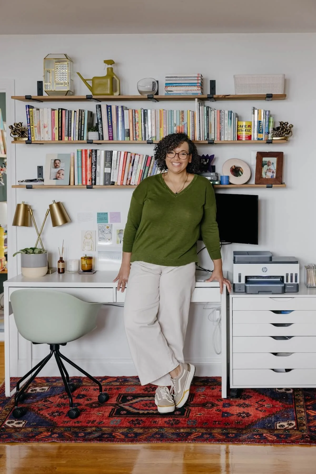 Brittney Nichols, a smiling woman with curly hair and glasses wearing a green sweater, beige pants, and sneakers, standing in a home office with bookshelves, a desk, a chair, and a colorful rug.