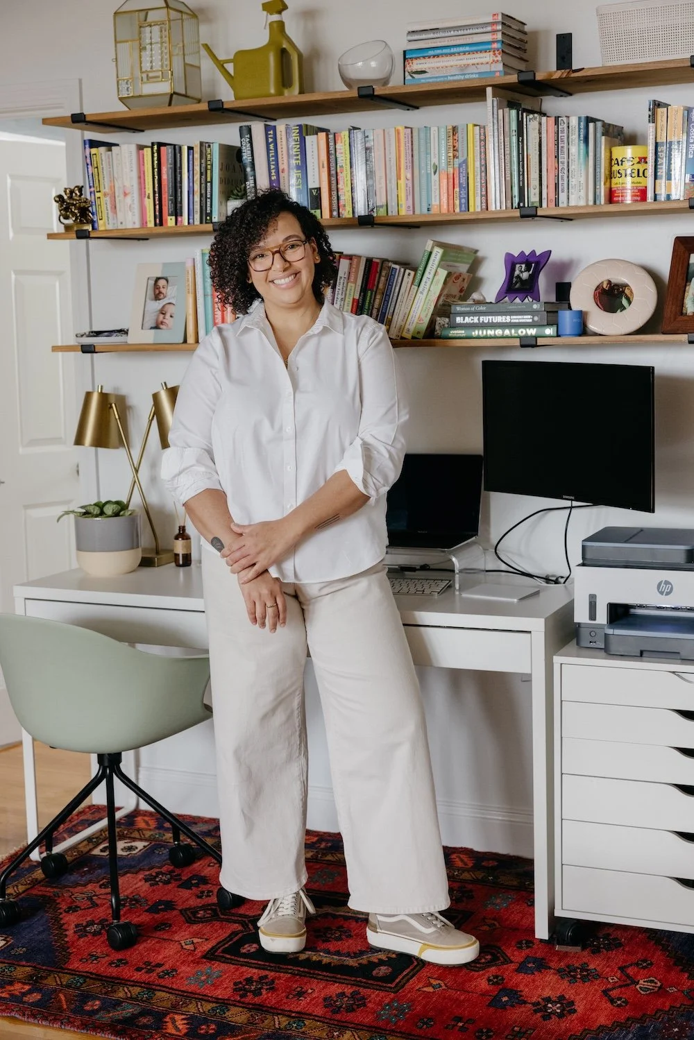 Brittney Nichols, a woman with curly hair and glasses smiling, standing in a home office with bookshelves, a desk with a computer and printer, and stylish decor.