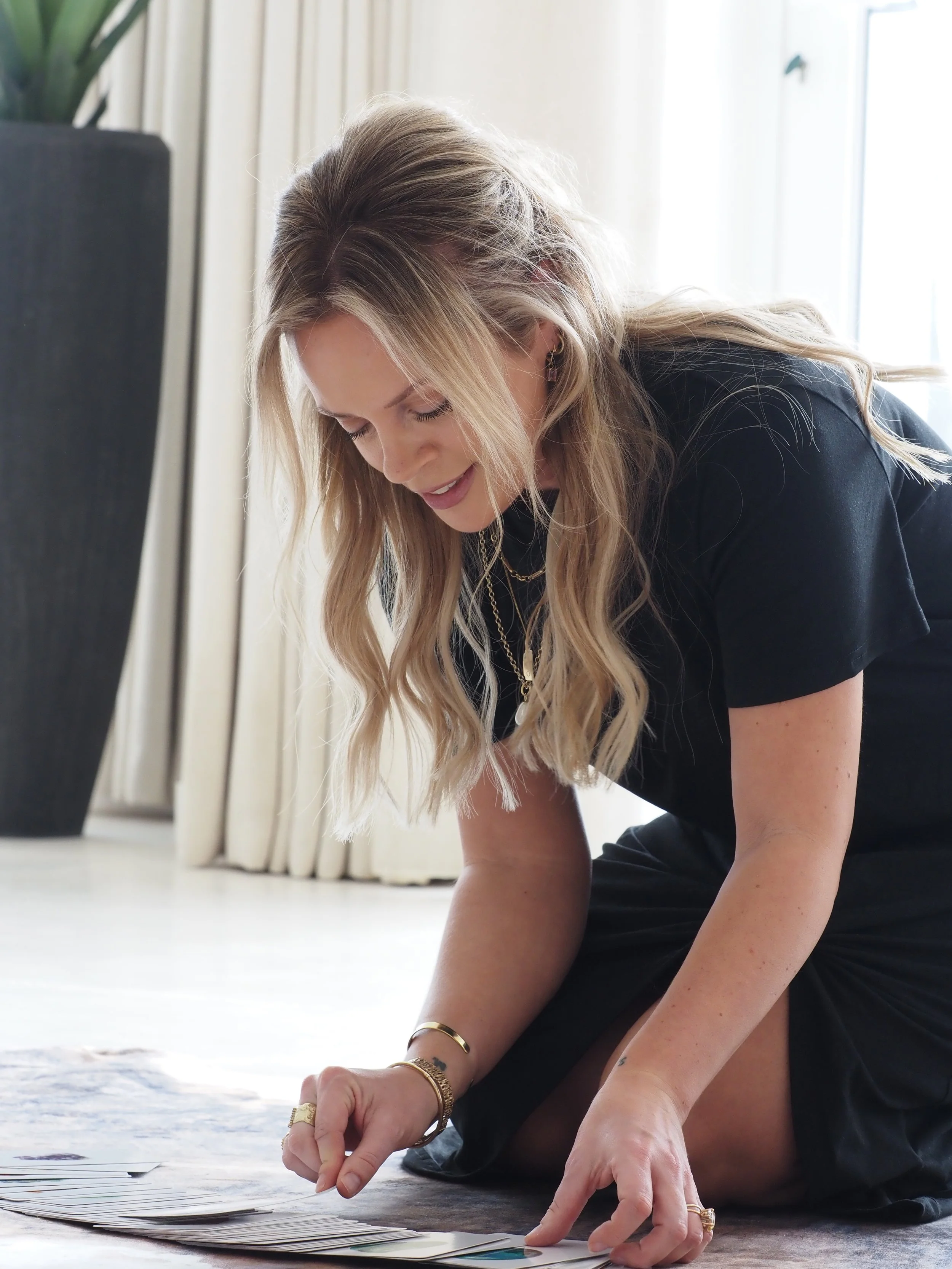 A woman with long blonde hair, wearing jewelry and a black shirt, is kneeling on the floor sorting through a stack of papers or photographs.