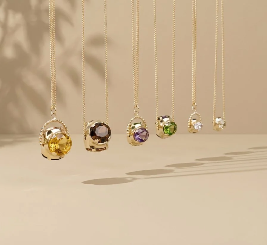 Six necklaces with colorful gemstone pendants hanging in a row against a beige background.