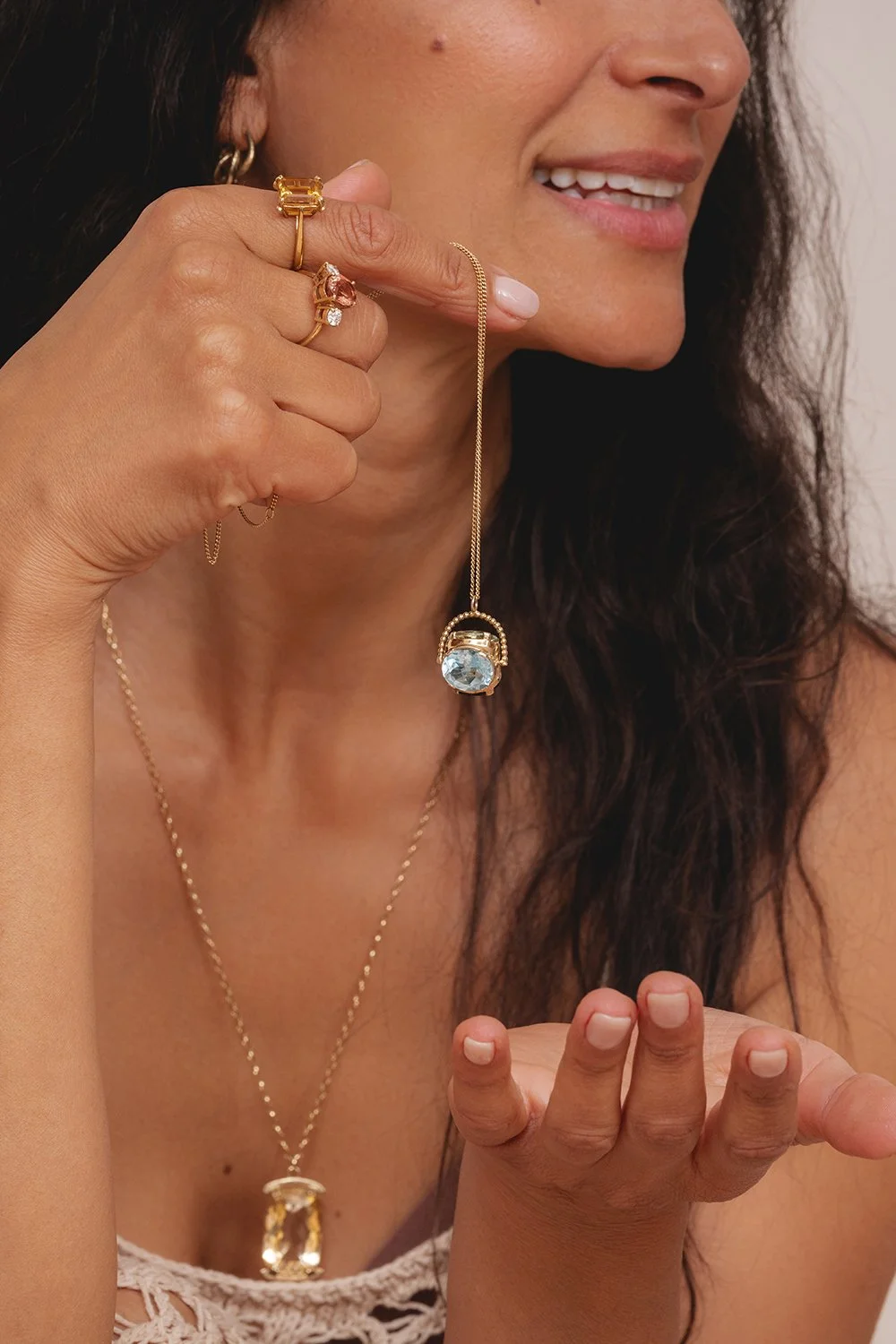 Close-up of a woman with dark, wavy hair, smiling slightly. She is wearing several gold jewelry pieces, including rings, a necklace with a large pendant, and a gold chain with a gemstone pendant. She is holding a delicate gold necklace with a large blue gemstone pendant near her face.