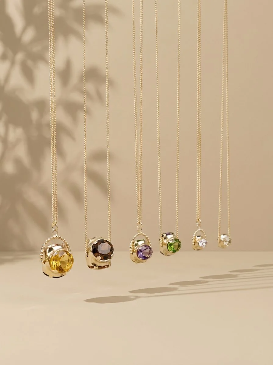 Six gold necklaces with pendants containing various colored gemstones, hanging against a light-colored background with shadows.
