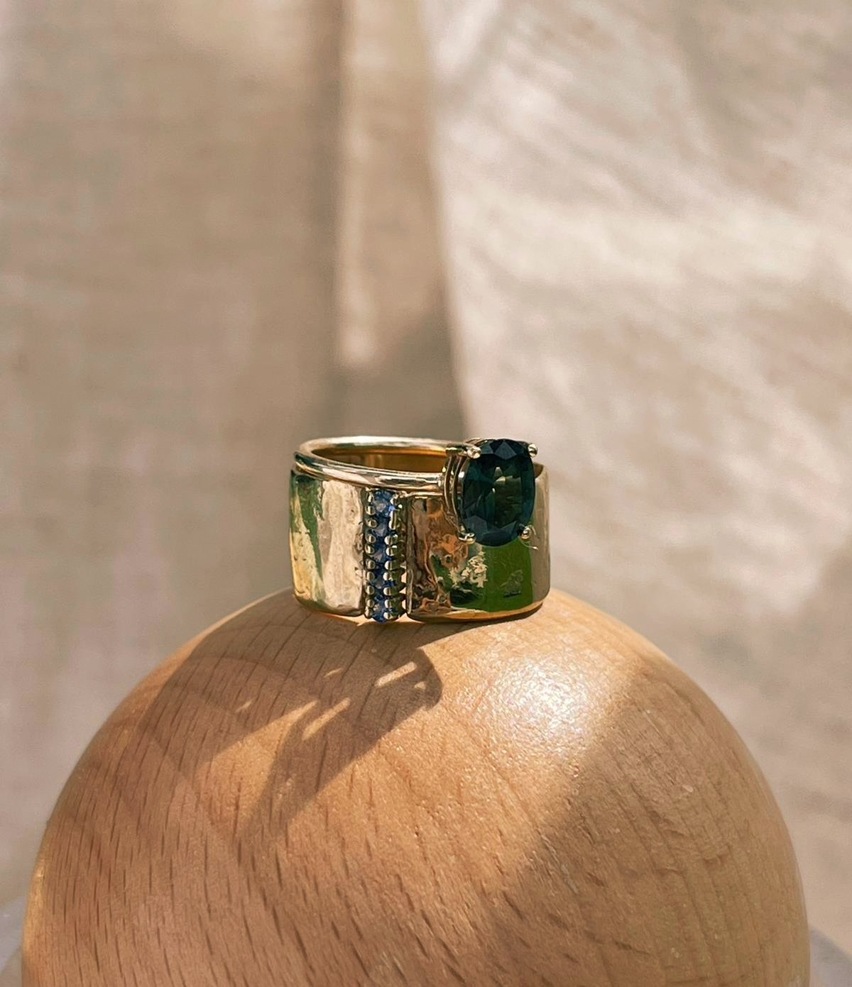 A gold ring with a dark green teal sapphire gemstone and small blue stones, placed on a wooden surface with a shadow, against a light background.