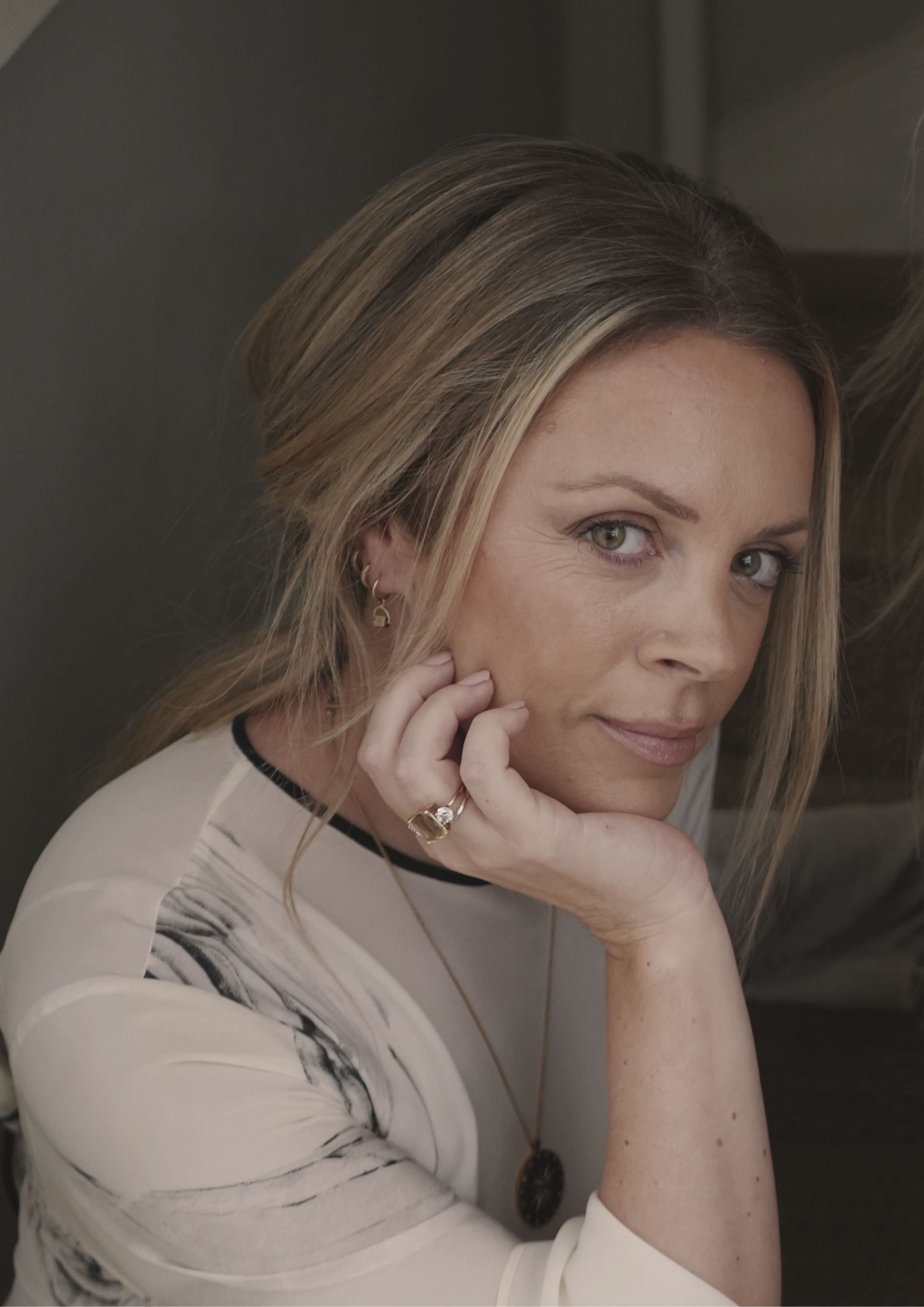 A woman with blonde hair, light skin, and green eyes resting her chin on her hand, looking at the camera, wearing earrings, a ring, and a necklace, dressed in a white top with black accents.