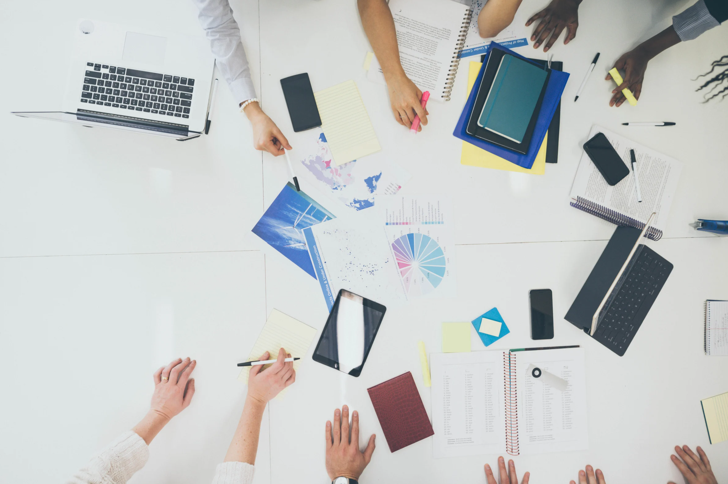 A top-down view of a group of people collaborating at a white table with laptops, tablets, smartphones, notebooks, pens, sticky notes, and printing materials, indicating a business strategy meeting.